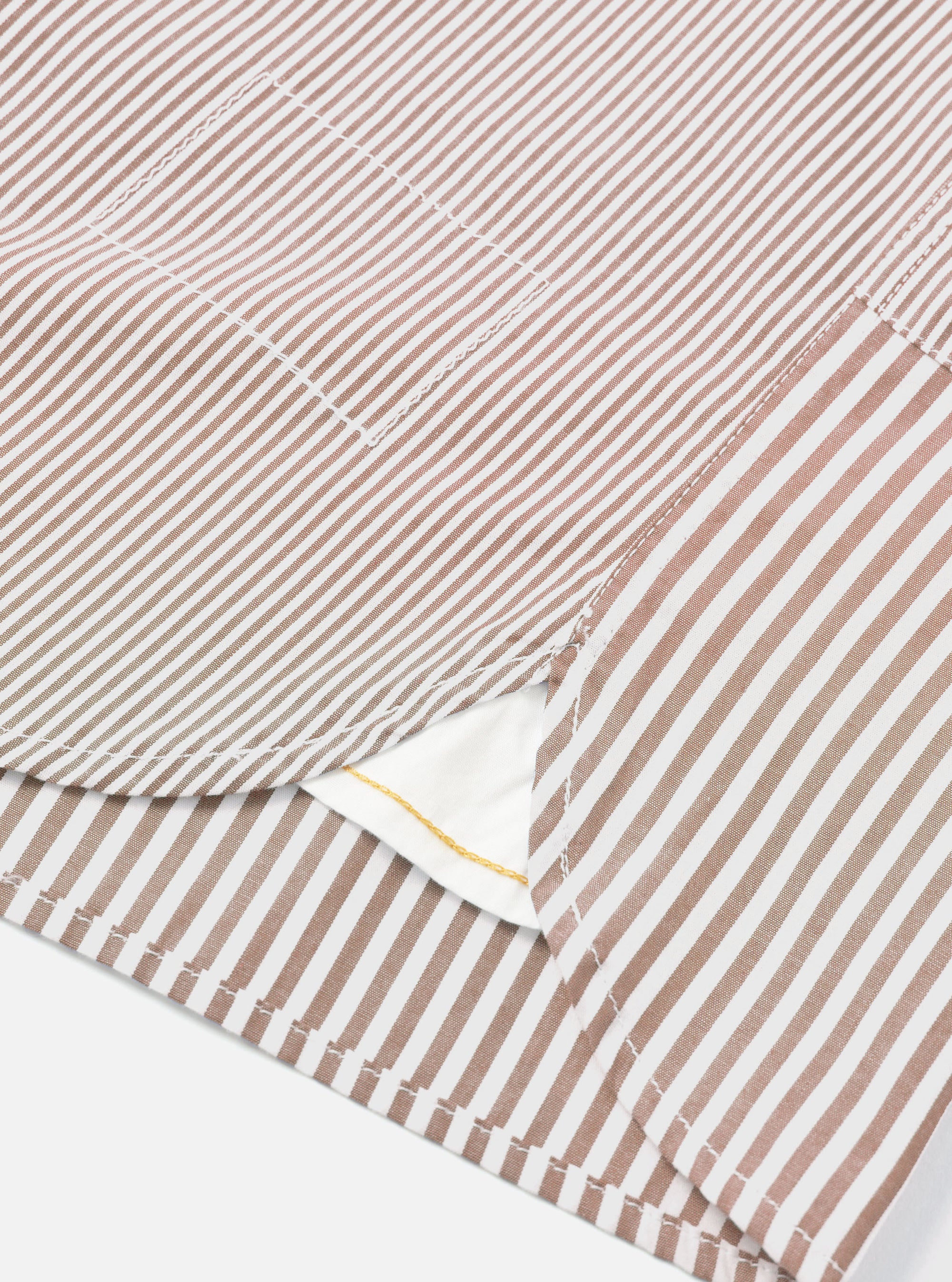 Close-up detail of a brown and white striped cotton shirt laid flat on a white surface. The shot focuses on the curved shirt tail hem, a chest patch pocket, and side seam stitching. A white inner label with a gold detail is visible at the hem opening.