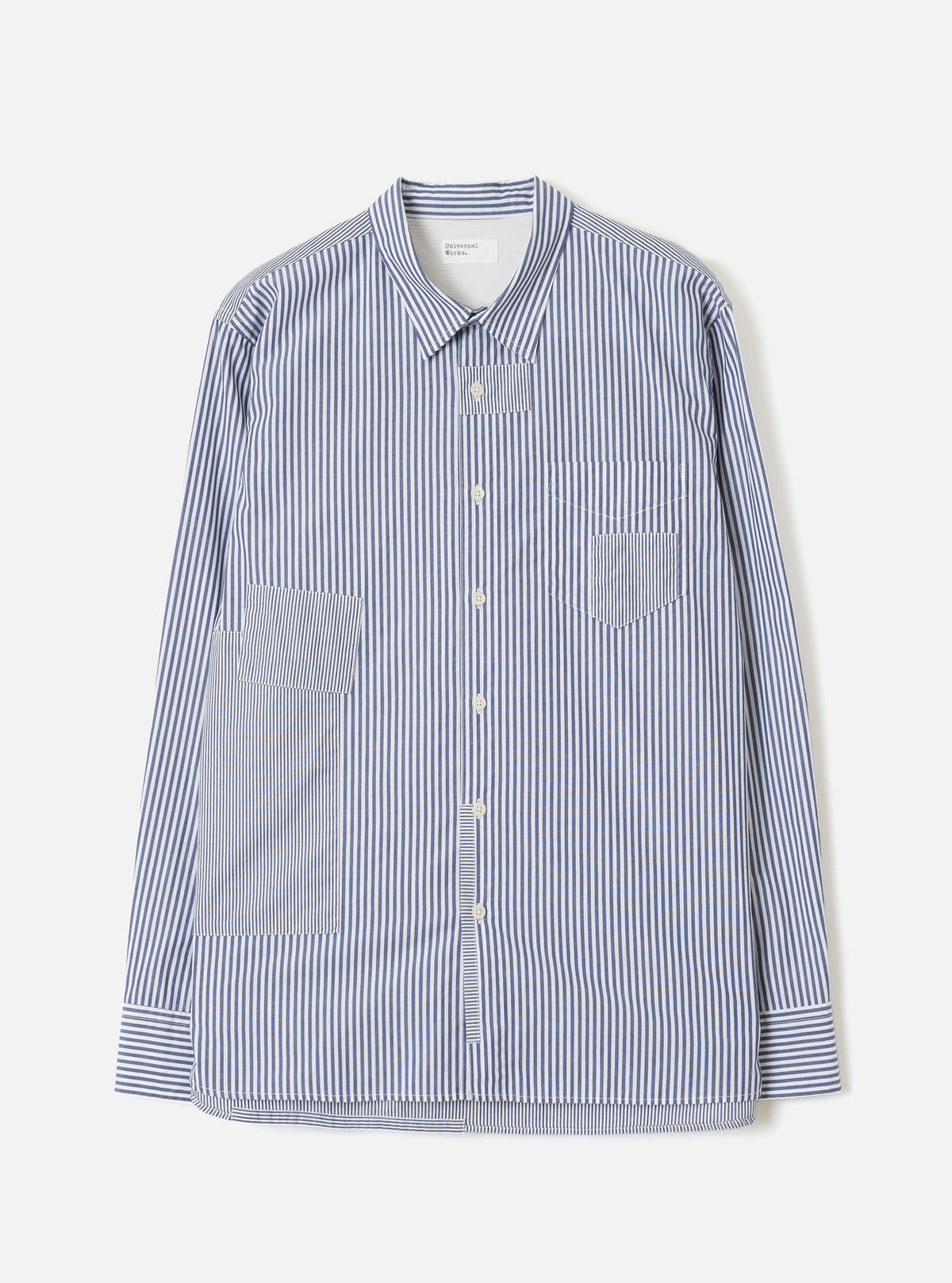 Navy and white vertical-stripe cotton shirt, flat lay on a light grey seamless backdrop. Spread collar, button-through placket with cream buttons, one chest patch pocket and two large front patch pockets. Long sleeves with buttoned cuffs. Topstitched hem with a curved silhouette. Universal Works label visible at the collar.