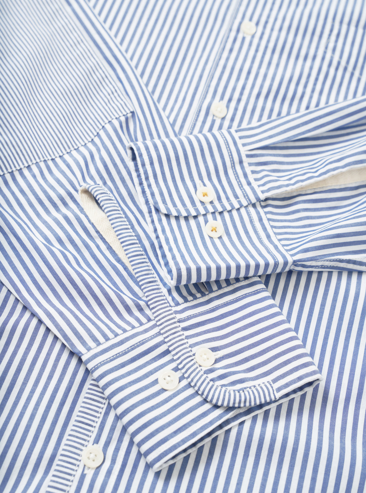 Close-up flat lay of a navy and white Bengal-stripe cotton shirt. Two barrel cuffs with white buttons are prominent, alongside a button placket with white buttons and a small chest patch pocket. The fabric has a crisp, smooth texture. No model or mannequin present.