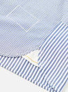 Close-up detail of a navy and white stripe cotton shirt laid flat on a white surface. The shot focuses on the curved hem, side seam vent, and a chest patch pocket. Topstitching runs along the hem and pocket edges. A white inner layer and small gold label are visible through the open vent.