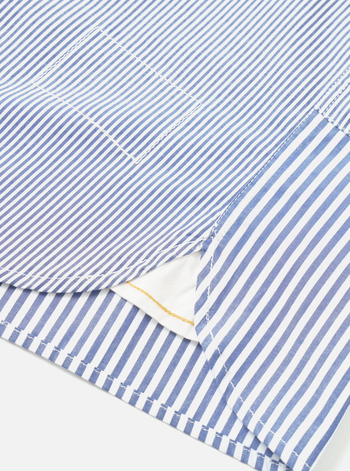 Close-up detail of a navy and white stripe cotton shirt laid flat on a white surface. The shot focuses on the curved hem, side seam vent, and a chest patch pocket. Topstitching runs along the hem and pocket edges. A white inner layer and small gold label are visible through the open vent.