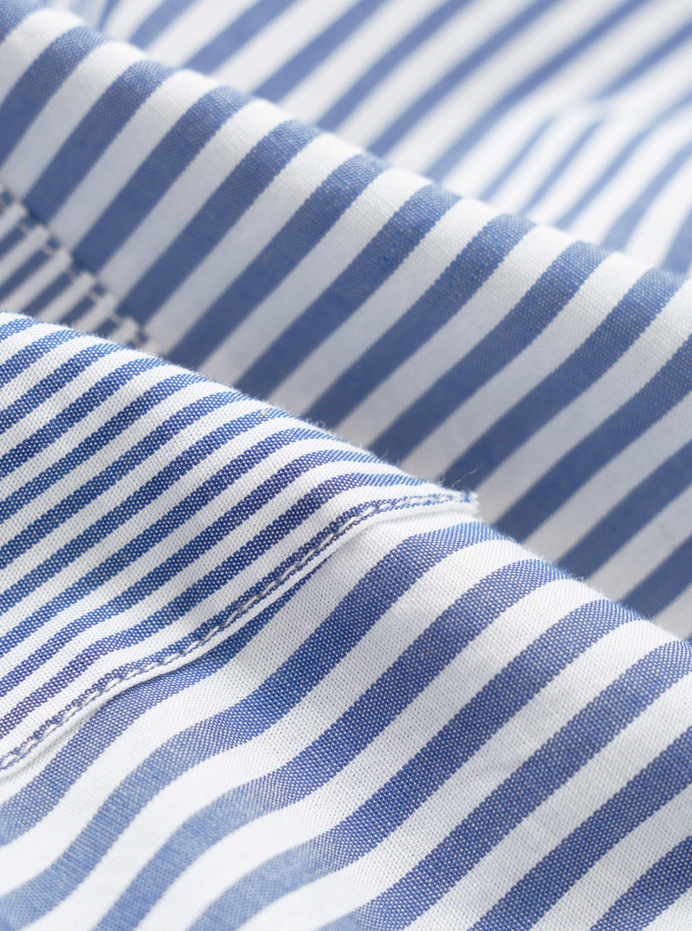Close-up detail of the navy and white Bengal stripe cotton fabric on the Queens patched shirt. The shot highlights the woven texture and a neat seam joining two panels, with fine topstitching visible along the edge.