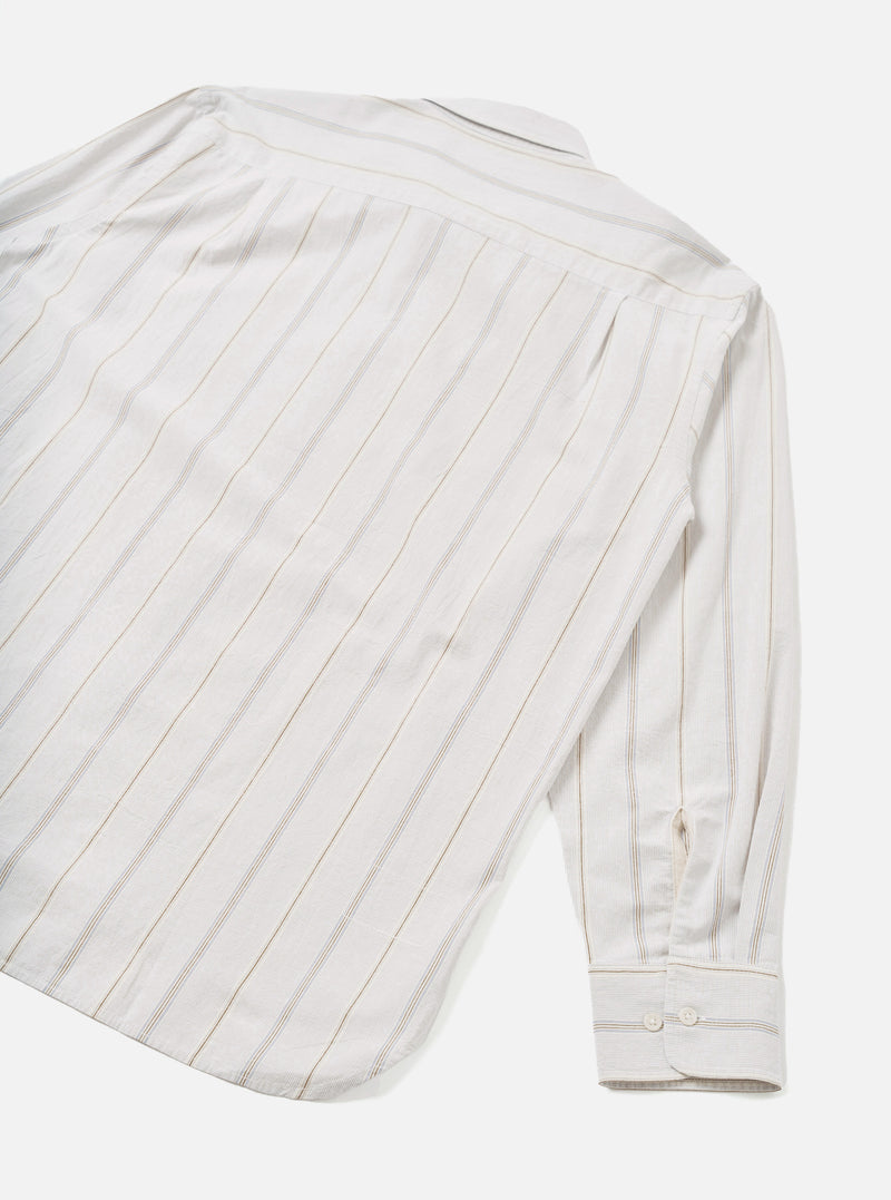 Flat lay of the Dove Monk Organic Stripe Square Pocket Shirt on a white studio backdrop, shot from above at an angle showing the back and one sleeve. Off-white woven fabric with vertical tan and grey pinstripes. Spread collar and buttoned cuff visible, with a curved hem at the base.