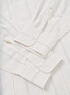 Close-up flat lay of the Dove Monk organic stripe square pocket shirt in off-white, focusing on the cuffs and front placket. The woven fabric shows fine vertical stripes in blue and tan. Barrel cuffs feature white buttons with gold bartack stitching. The front placket displays white buttons on a plain white surface.