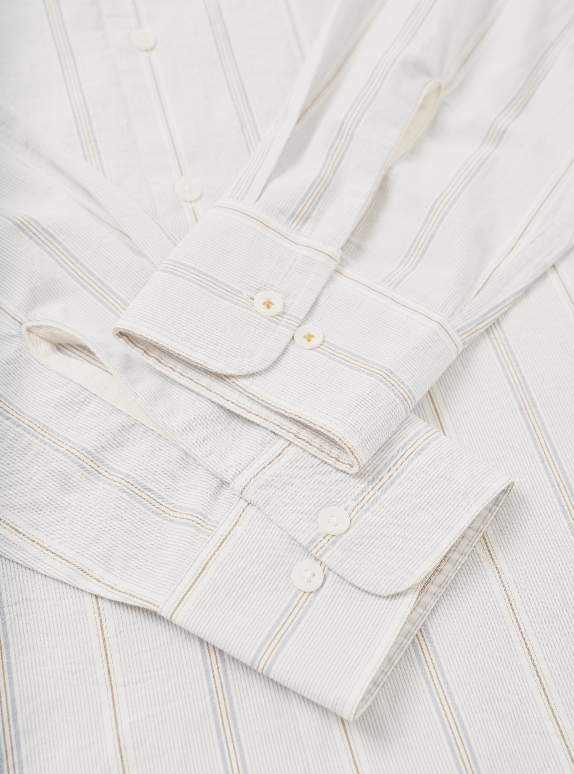 Close-up flat lay of the Dove Monk organic stripe square pocket shirt in off-white, focusing on the cuffs and front placket. The woven fabric shows fine vertical stripes in blue and tan. Barrel cuffs feature white buttons with gold bartack stitching. The front placket displays white buttons on a plain white surface.