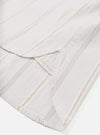 Close-up flat lay of the hem and side vent detail on a dove white organic stripe square-pocket shirt. The woven fabric features vertical stripes in grey, tan, and a fine gold thread on a white ground. A curved side vent opening is visible, showing the clean finished hem and inner facing. Shot on a white studio backdrop.