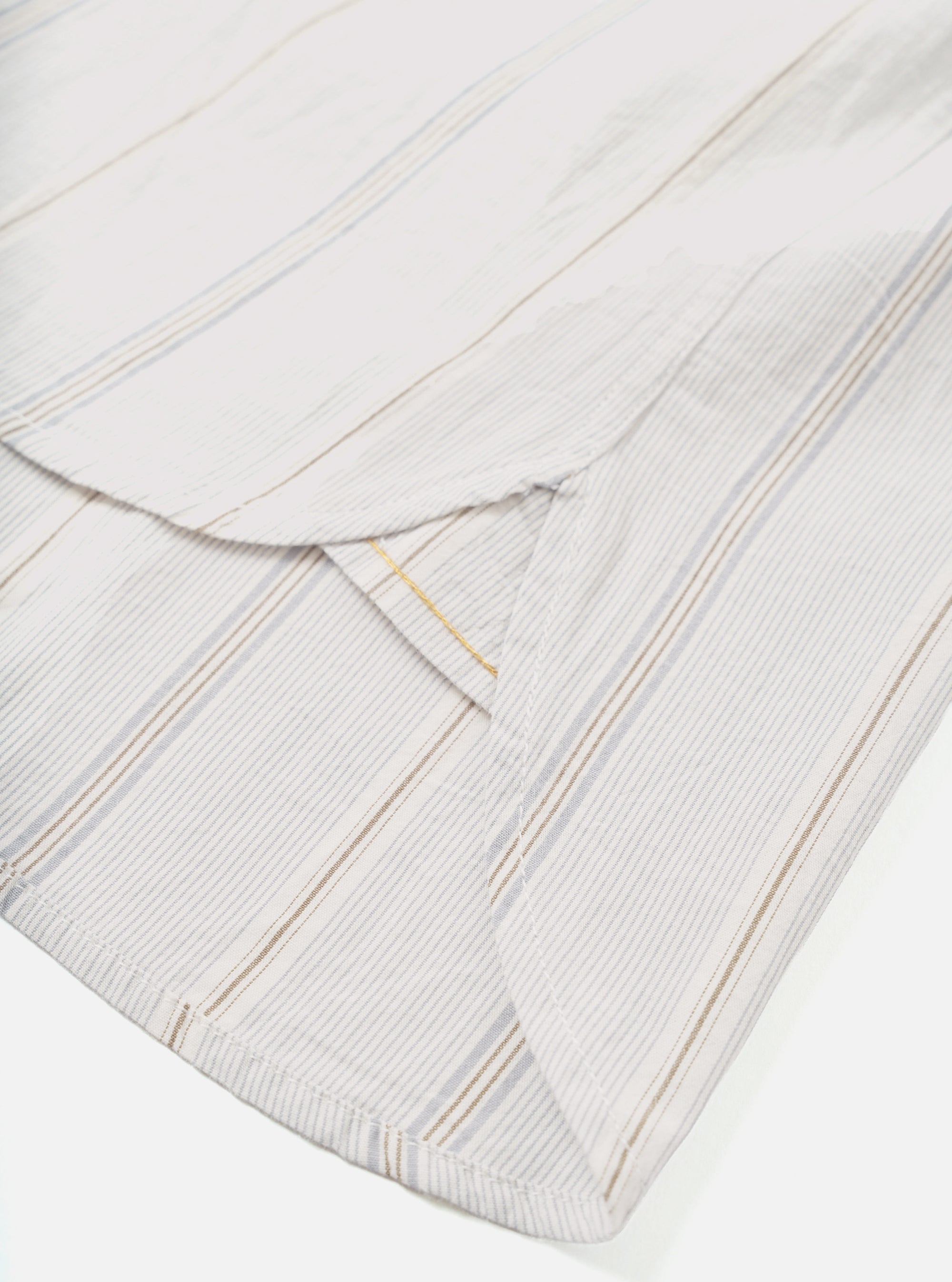 Close-up flat lay of the hem and side vent detail on a dove white organic stripe square-pocket shirt. The woven fabric features vertical stripes in grey, tan, and a fine gold thread on a white ground. A curved side vent opening is visible, showing the clean finished hem and inner facing. Shot on a white studio backdrop.