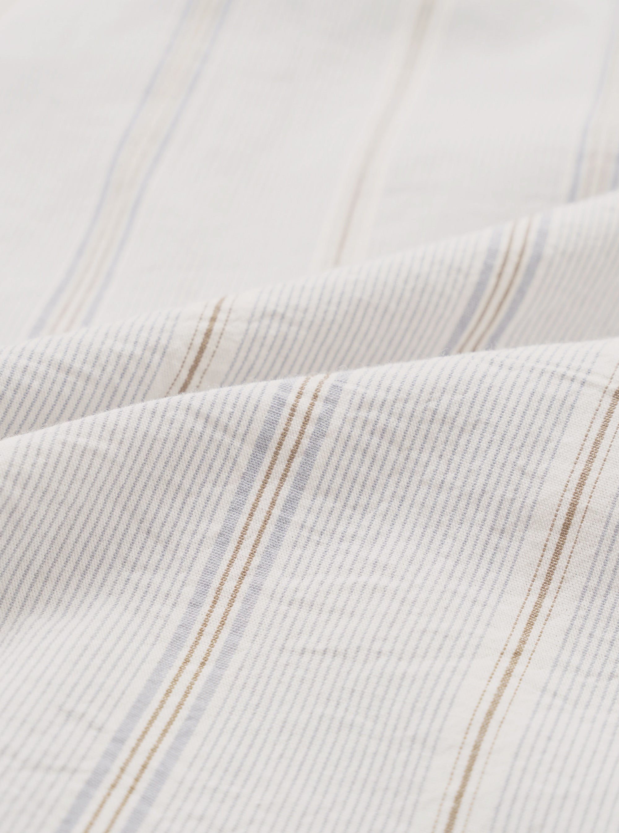 Close-up of the organic stripe fabric on the Dove Monk pocket shirt. The off-white woven cloth features fine vertical pinstripes in pale blue and tan, grouped into repeat stripe clusters. The soft, lightweight texture is visible across two folded layers of fabric against a white studio backdrop.