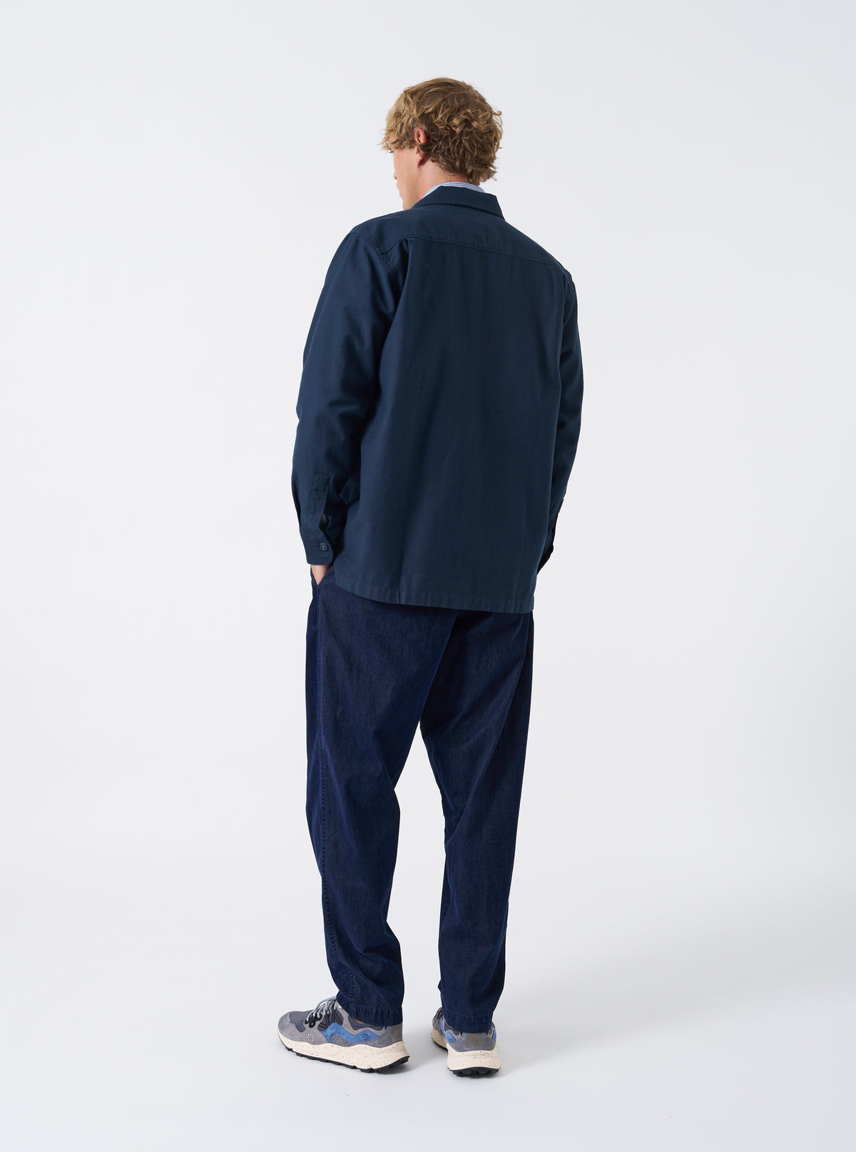A male model wearing the Navy Slub Sateen Fatigue Shirt is captured from the back against a plain white background. The shot displays the relaxed shoulder fit and casual length of the garment.