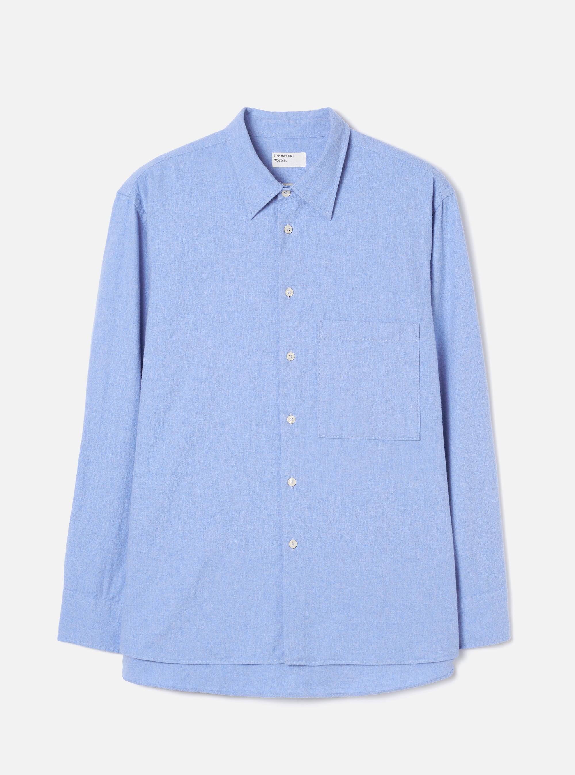 Flat lay of a blue brushed flannel shirt on a light grey studio backdrop. Features a pointed collar, full-length sleeves with plain cuffs, a buttoned placket with six small white buttons, and a square patch pocket on the left chest. The curved shirttail hem is visible at the front. Fabric has a soft, lightly textured finish.