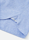 Close-up of the hem and side seam on a blue brushed flannel shirt, shot on a white studio backdrop. A curved side vent is visible with a small yellow bar tack stitch at its base. The Oxford-weave flannel fabric shows a soft, slightly mottled texture in a mid-blue chambray tone.