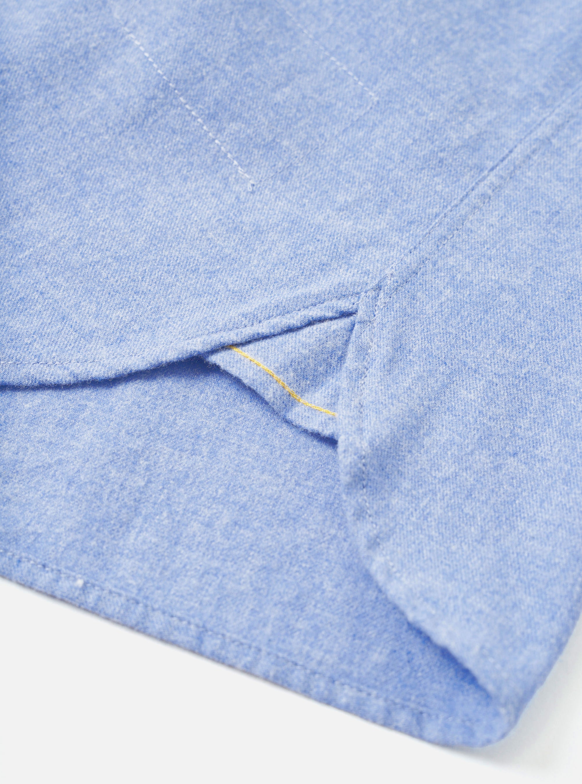 Close-up of the hem and side seam on a blue brushed flannel shirt, shot on a white studio backdrop. A curved side vent is visible with a small yellow bar tack stitch at its base. The Oxford-weave flannel fabric shows a soft, slightly mottled texture in a mid-blue chambray tone.
