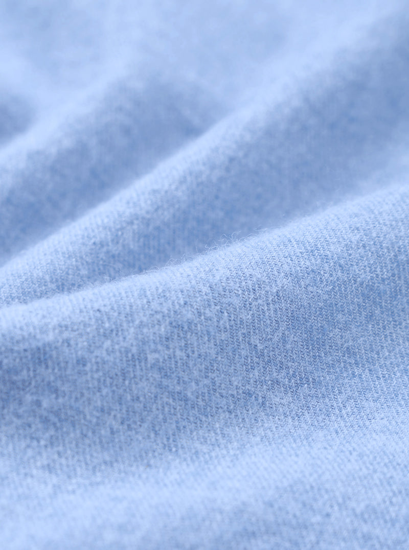 Close-up of the brushed flannel fabric used in the blue square pocket shirt. The tight knit weave is clearly visible, with a soft, lightly napped surface in a pale sky blue. Gentle folds show the fabric's drape and texture across the full frame.