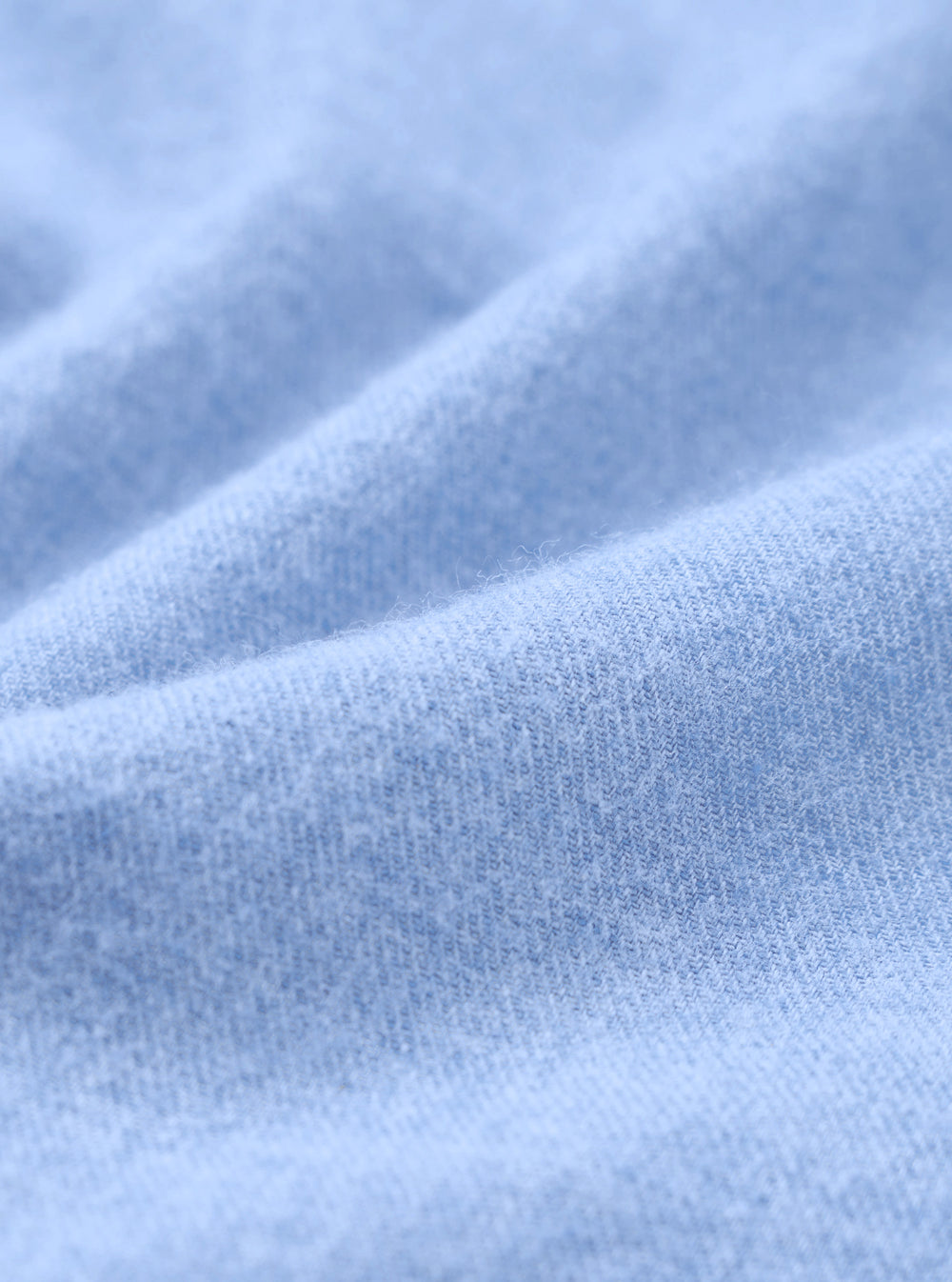 Close-up of the brushed flannel fabric used in the blue square pocket shirt. The tight knit weave is clearly visible, with a soft, lightly napped surface in a pale sky blue. Gentle folds show the fabric's drape and texture across the full frame.