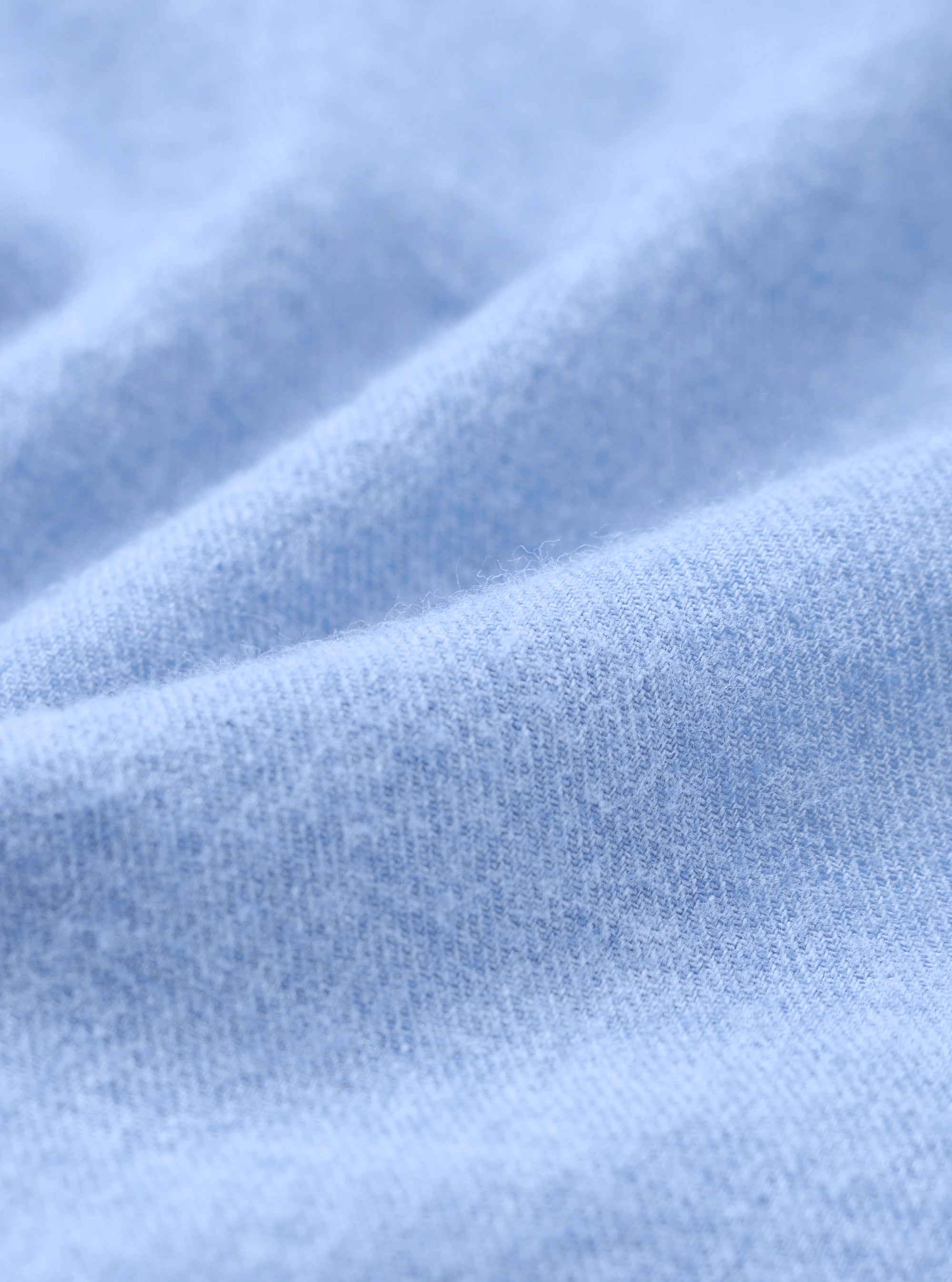 Close-up of the brushed flannel fabric used in the blue square pocket shirt. The tight knit weave is clearly visible, with a soft, lightly napped surface in a pale sky blue. Gentle folds show the fabric's drape and texture across the full frame.
