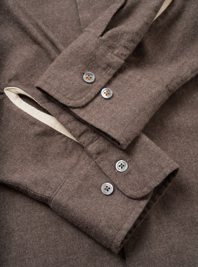 Close-up flat lay of the cuffs of a brown brushed flannel shirt. Each barrel cuff fastens with two four-hole grey buttons. The flannel has a soft, lightly textured mélange weave. A contrast natural ecru lining is visible at the cuff opening. Neat topstitching runs along the cuff edges.