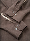 Close-up flat lay of the cuffs of a brown brushed flannel shirt. Each barrel cuff fastens with two four-hole grey buttons. The flannel has a soft, lightly textured mélange weave. A contrast natural ecru lining is visible at the cuff opening. Neat topstitching runs along the cuff edges.