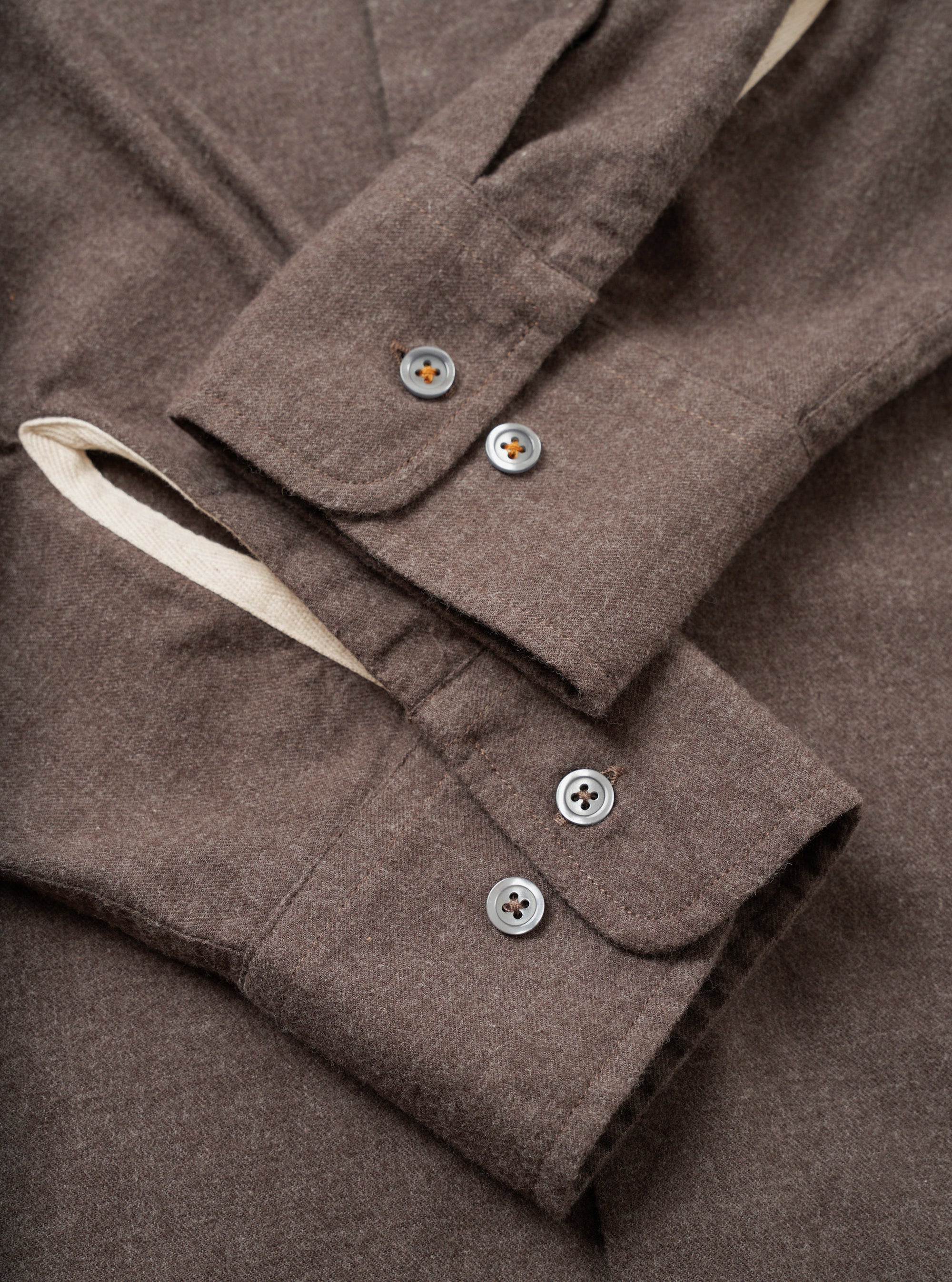 Close-up flat lay of the cuffs of a brown brushed flannel shirt. Each barrel cuff fastens with two four-hole grey buttons. The flannel has a soft, lightly textured mélange weave. A contrast natural ecru lining is visible at the cuff opening. Neat topstitching runs along the cuff edges.