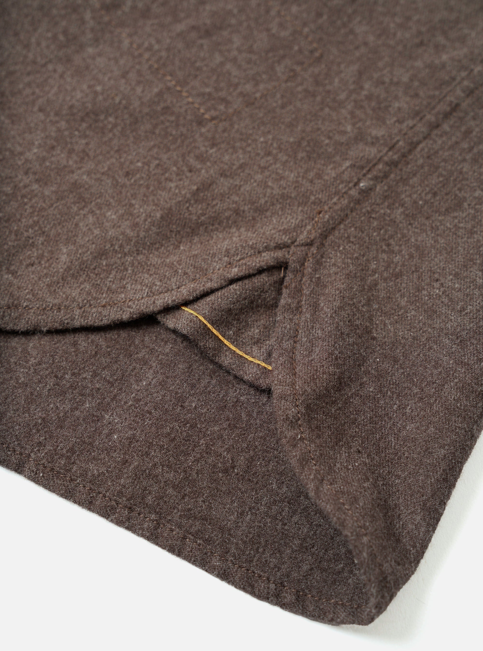 Close-up detail of a brown brushed flannel shirt laid flat on a white surface. The shot focuses on the curved shirt hem and a side seam vent, with a yellow contrast stitch visible along the vent opening. A small button is visible at the upper right. The fabric shows a soft, mélange flannel texture.
