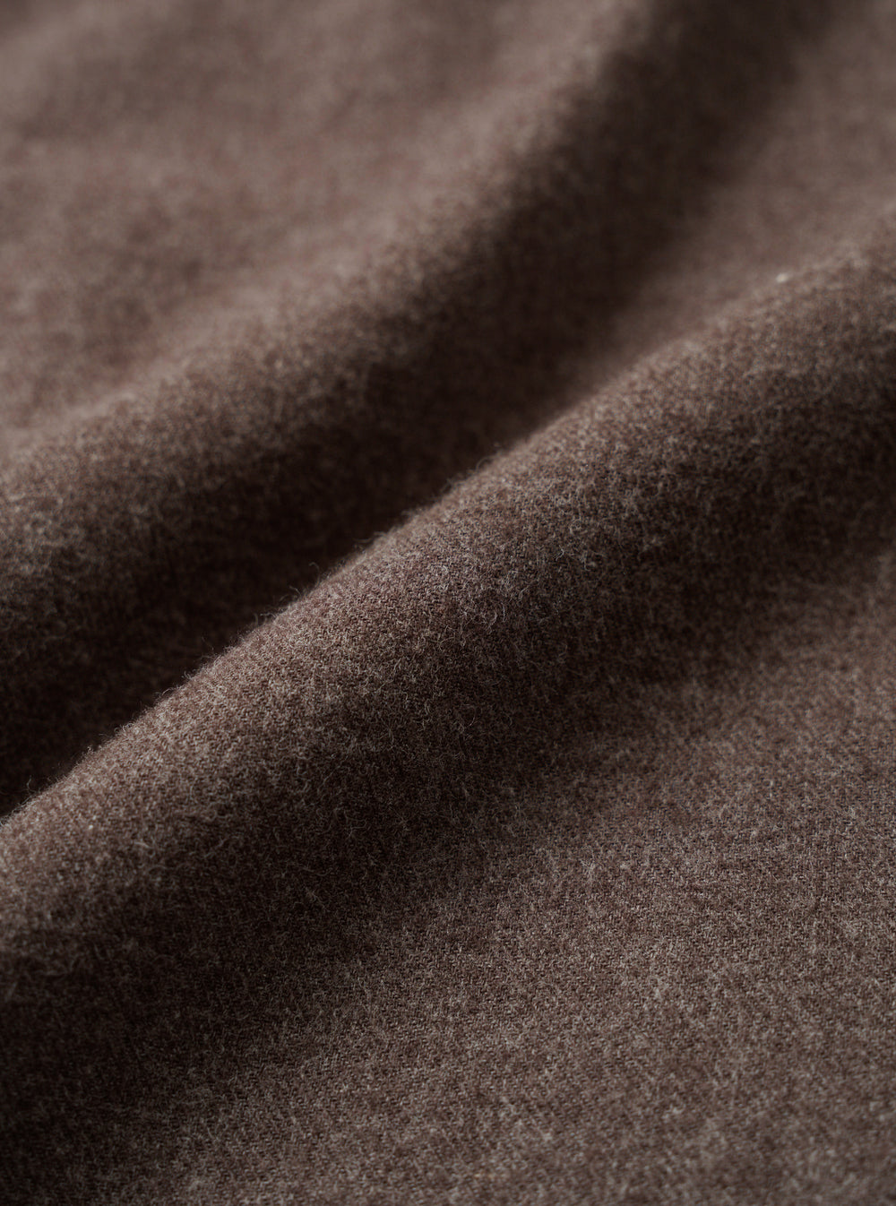 Close-up detail shot of the brown brushed flannel fabric used in the square pocket shirt. The textile shows a soft, lightly napped surface with fine fibres visible across the weave. The cloth is folded to reveal its drape and depth of colour, a warm muted brown with subtle grey undertones.