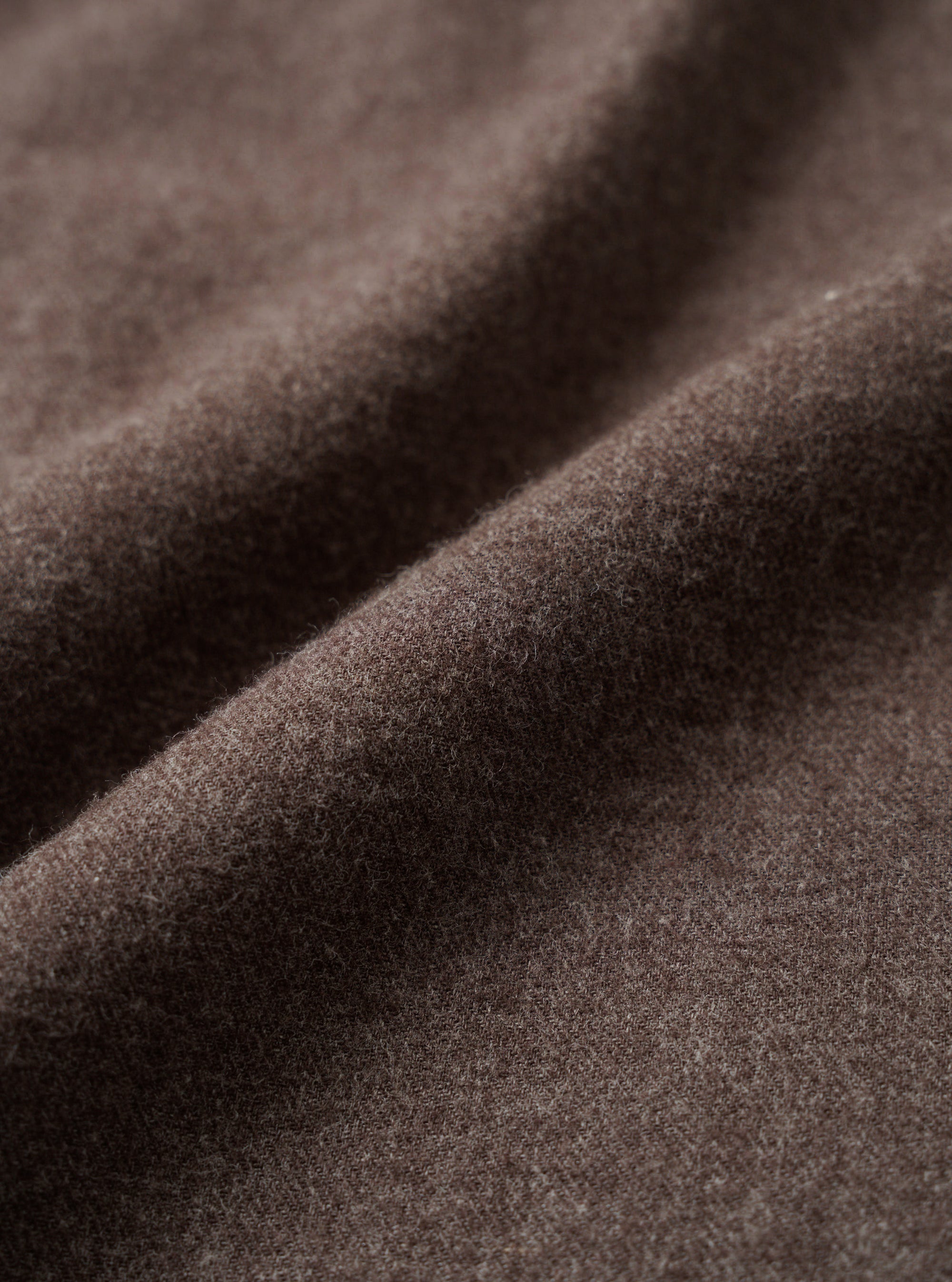 Close-up detail shot of the brown brushed flannel fabric used in the square pocket shirt. The textile shows a soft, lightly napped surface with fine fibres visible across the weave. The cloth is folded to reveal its drape and depth of colour, a warm muted brown with subtle grey undertones.