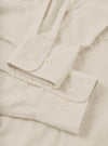 Close-up flat lay of an ecru brushed flannel shirt, focusing on two rounded cuffs fastened with white four-hole buttons and small gold cross-stitch details. The placket and a square chest pocket are visible above. The fabric has a soft, lightly textured surface. Shot on a plain ecru surface.