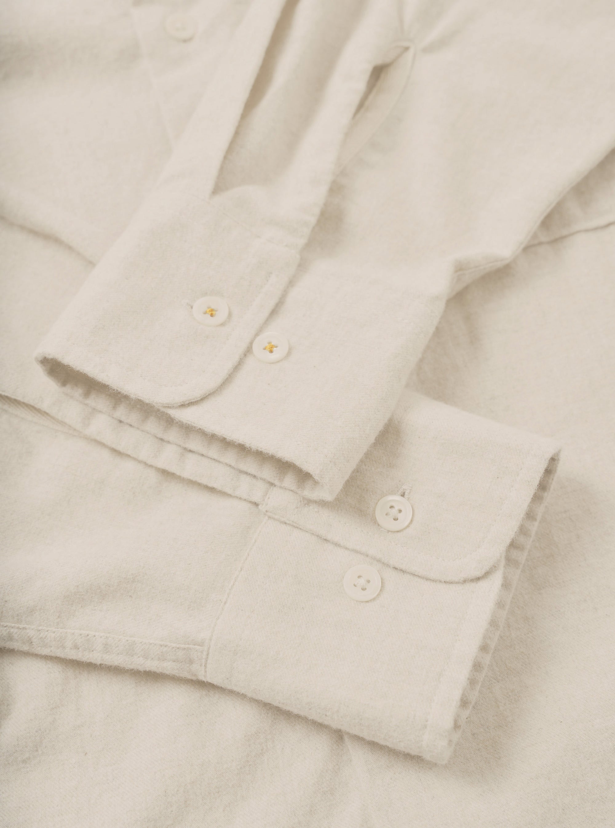 Close-up flat lay of an ecru brushed flannel shirt, focusing on two rounded cuffs fastened with white four-hole buttons and small gold cross-stitch details. The placket and a square chest pocket are visible above. The fabric has a soft, lightly textured surface. Shot on a plain ecru surface.