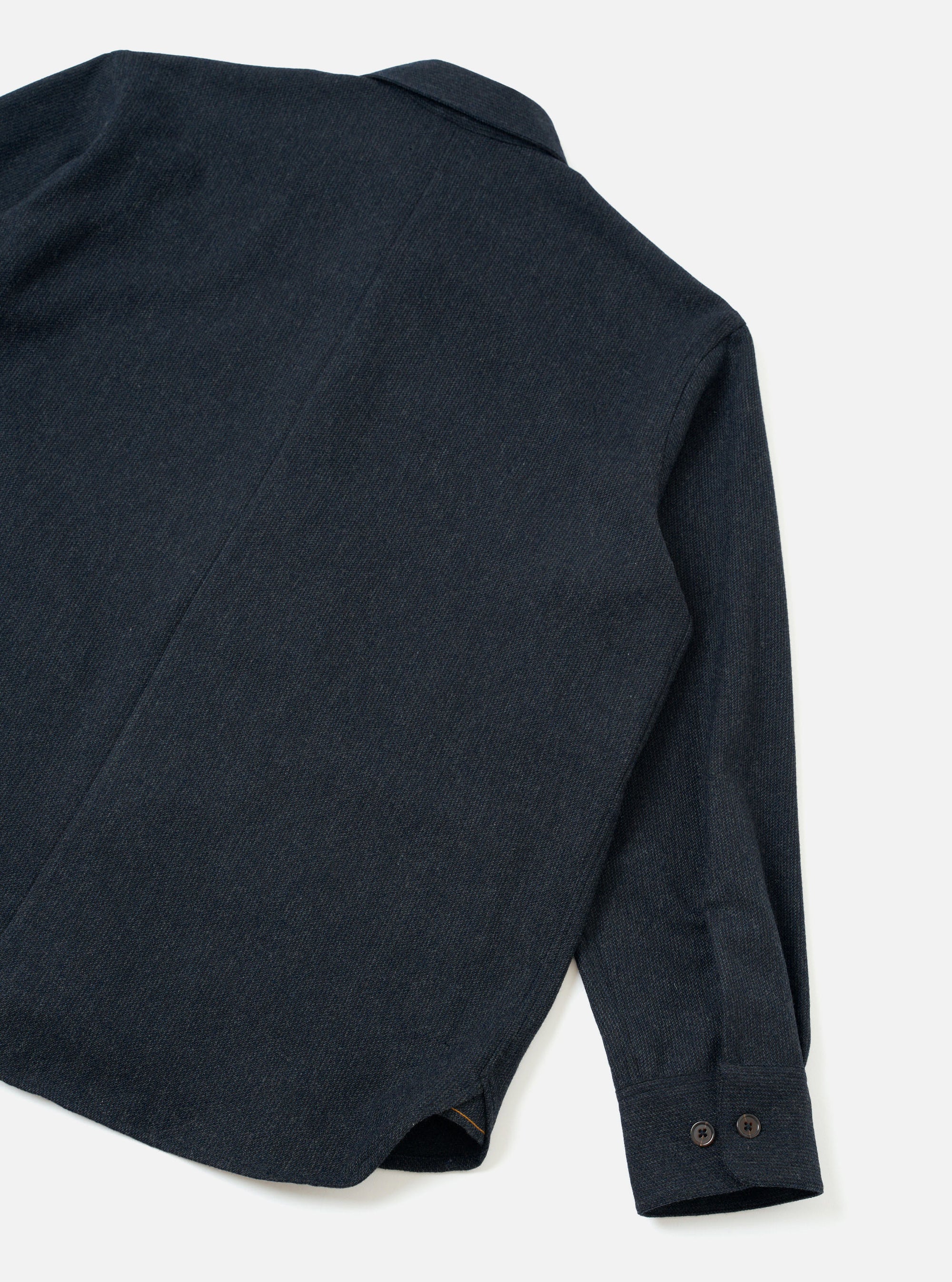 Flat lay of a navy flannel overshirt on a white surface, shot from above at an angle showing the back and right sleeve. The textured twill-weave flannel fabric has a subtle melange finish. A seam runs vertically down the back. The cuff is fastened with two dark horn-effect buttons. The curved shirt-tail hem is visible at the bottom corner.
