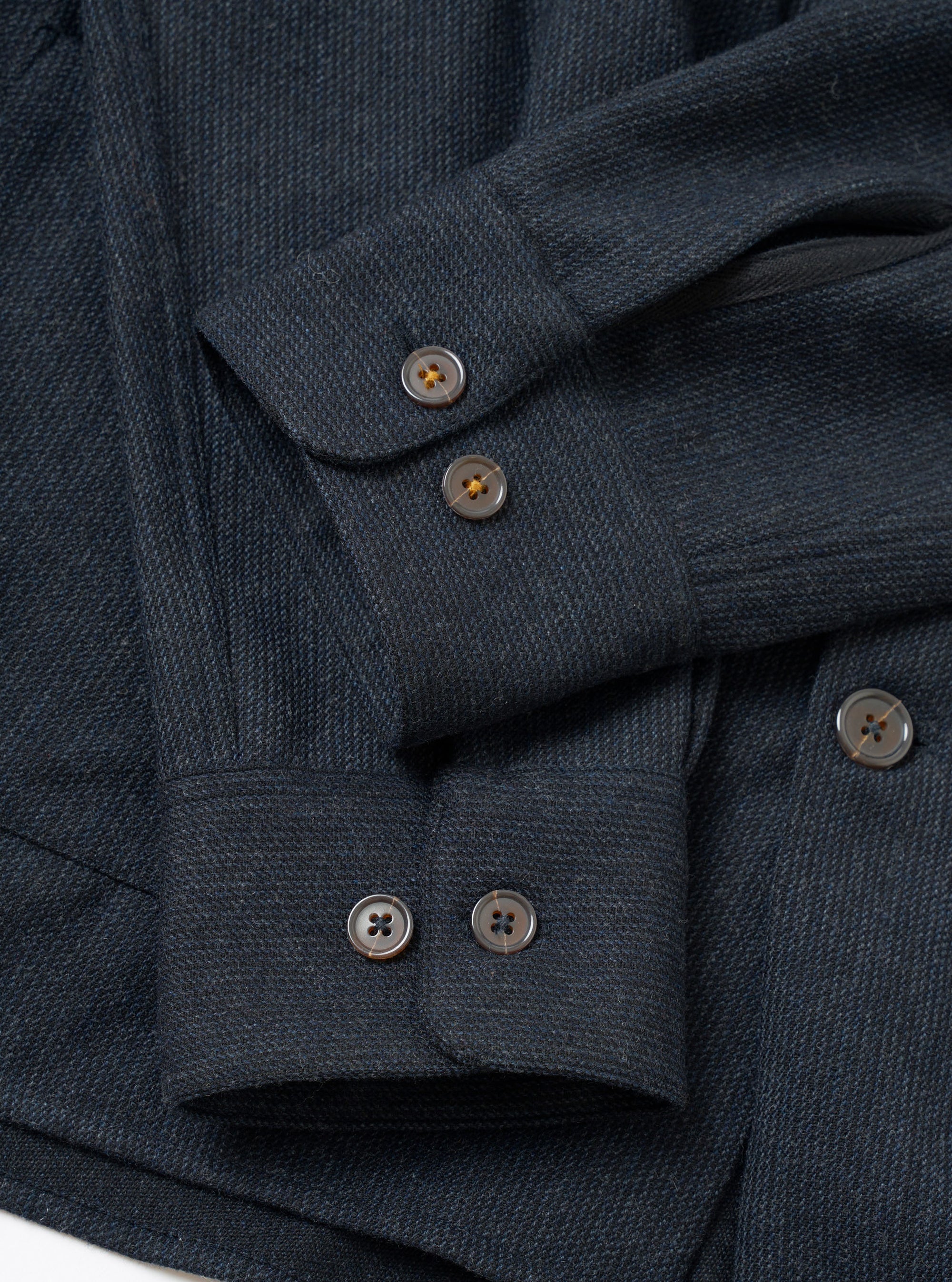 Close-up flat lay of a navy flannel overshirt on a white surface, focusing on the cuffs and front placket. The fabric has a textured twill weave with subtle tonal stripe. Four-hole horn-effect buttons fasten the barrel cuffs and placket. Two overlapping sleeves show double-button cuffs in detail.