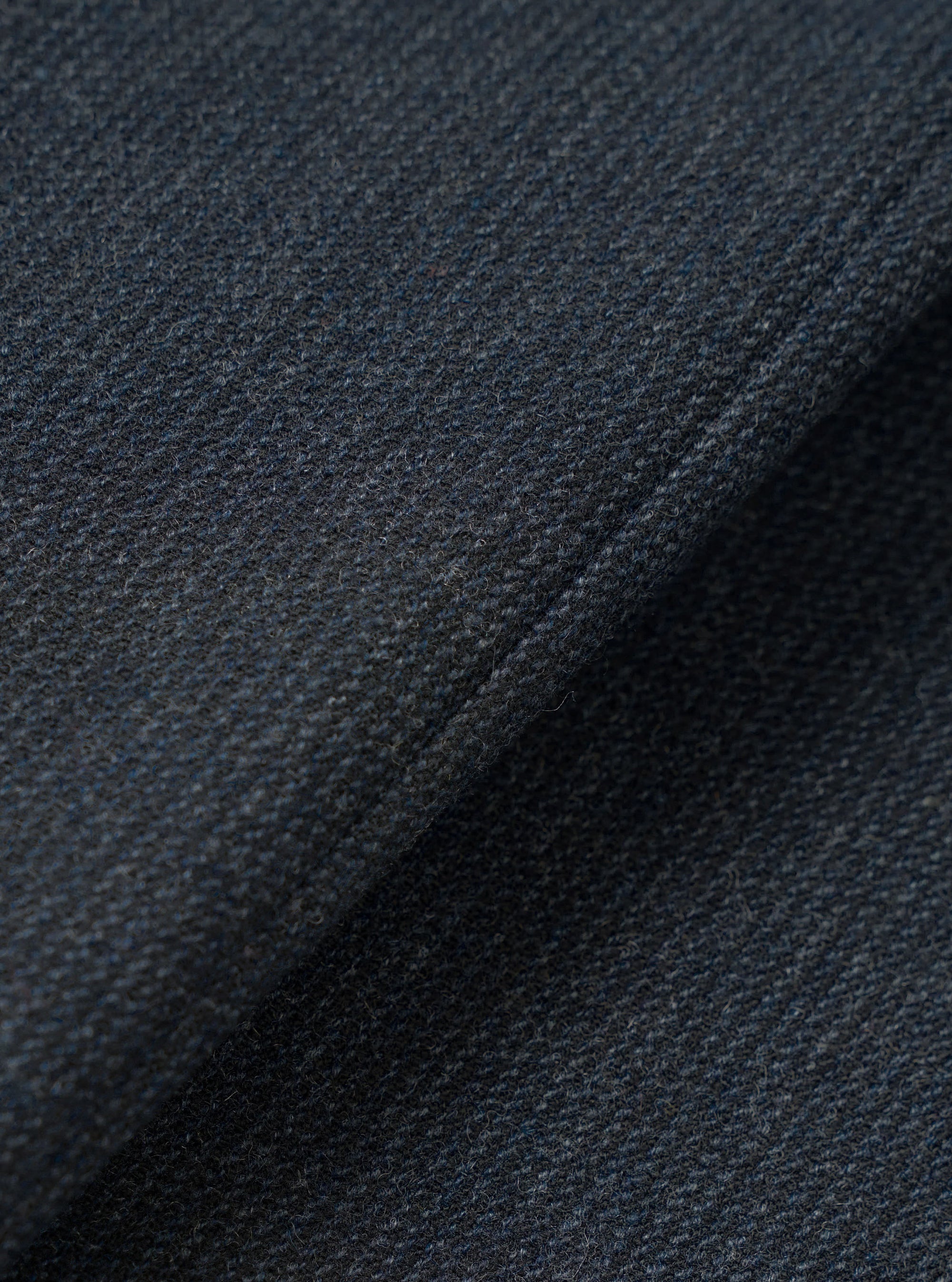 Close-up of the navy flannel fabric used in the Airline Travail Overshirt. The tight, nubby weave shows a dark navy base with flecks of grey and blue throughout, giving a heathered, textured appearance. A diagonal fold runs across the frame, highlighting the fabric's soft, lightly brushed surface.