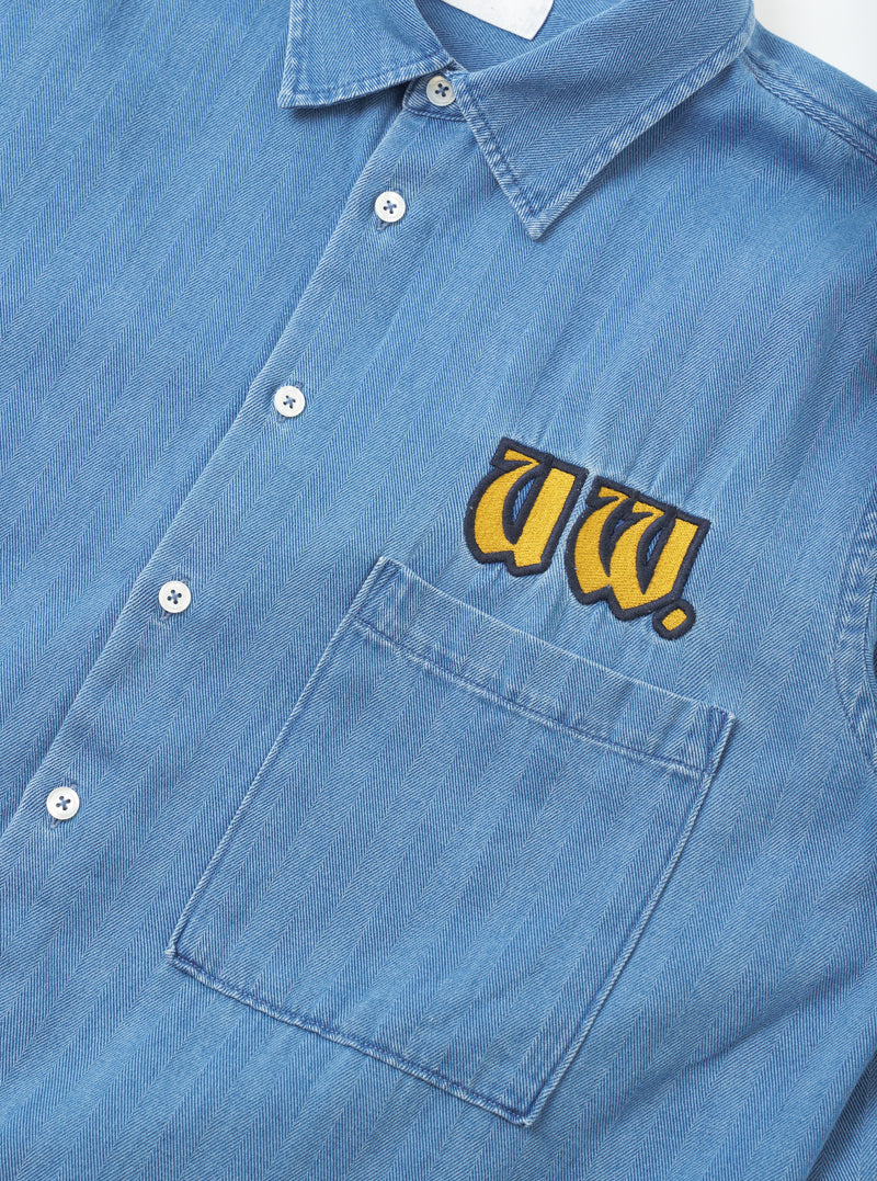 Close-up detail shot of the Washed Indigo Recycled Herringbone Patch Lucky Shirt on a plain background. The view focuses on the woven recycled cotton fabric, clean stitching, and classic button placket detailing.