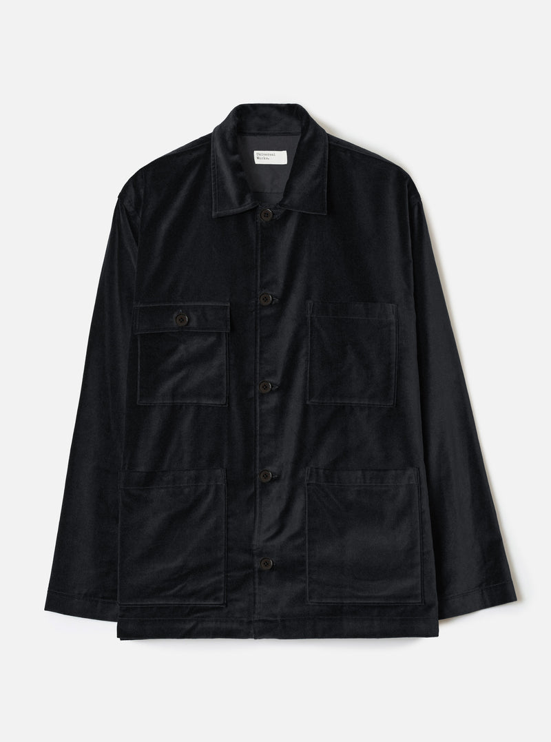 Black velvet chore jacket laid flat on a white studio backdrop. Spread collar with a Universal Works woven label at the neckline. Five dark buttons down the centre front placket. Two flap chest pockets and two large open patch pockets at the lower front. Long sleeves with plain cuffs. Straight hem.