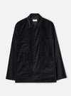 Black velvet chore jacket laid flat on a white studio backdrop. Spread collar with a Universal Works woven label at the neckline. Five dark buttons down the centre front placket. Two flap chest pockets and two large open patch pockets at the lower front. Long sleeves with plain cuffs. Straight hem.
