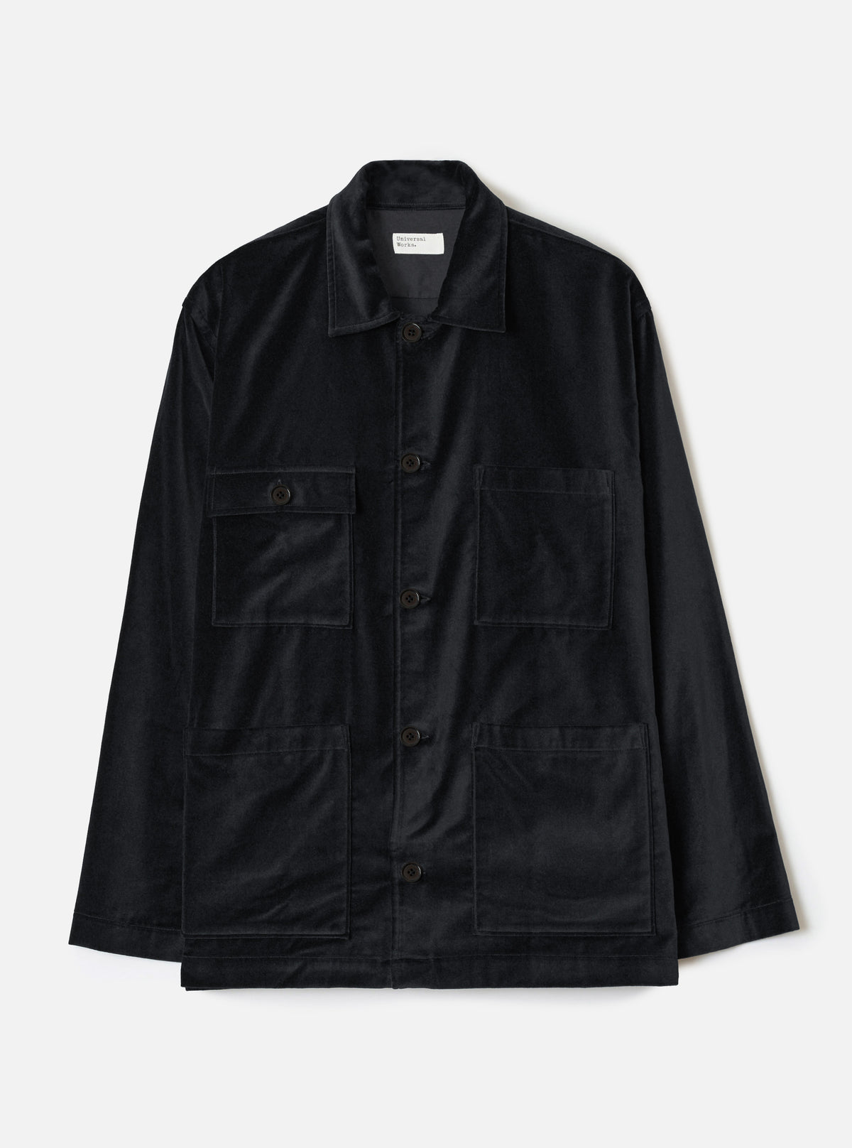Black velvet chore jacket laid flat on a white studio backdrop. Spread collar with a Universal Works woven label at the neckline. Five dark buttons down the centre front placket. Two flap chest pockets and two large open patch pockets at the lower front. Long sleeves with plain cuffs. Straight hem.