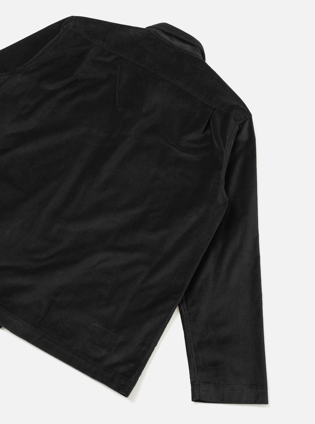 Black velvet dockside jacket shot flat on a light grey seamless backdrop, angled detail view showing the rear shoulder seam, full-length sleeve with a plain cuff, and a rear patch pocket at the lower left. The fabric has a soft, low-pile velvet texture with a subtle sheen. No model or mannequin present.