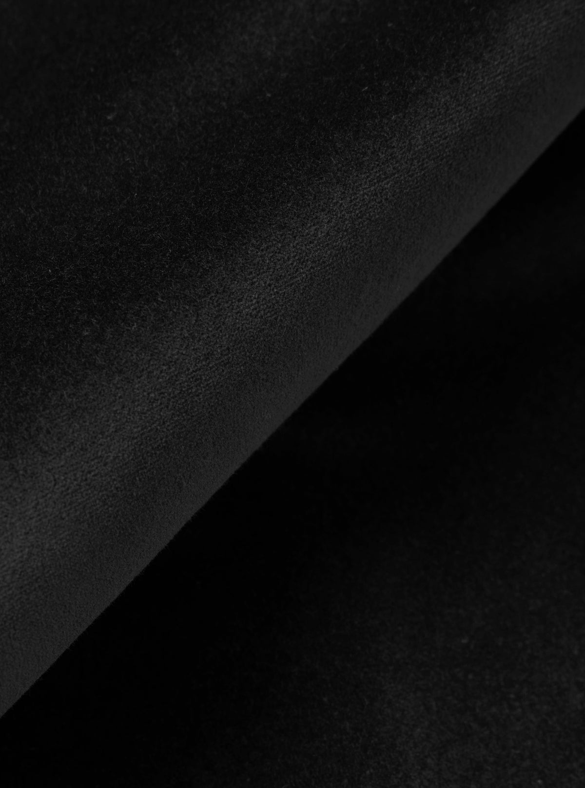 Close-up detail shot of black velvet fabric, showing a diagonal fold across the frame. The matte, fine-pile surface catches subtle light variation, revealing the texture and depth of the material against the darker shadow of the fold beneath.