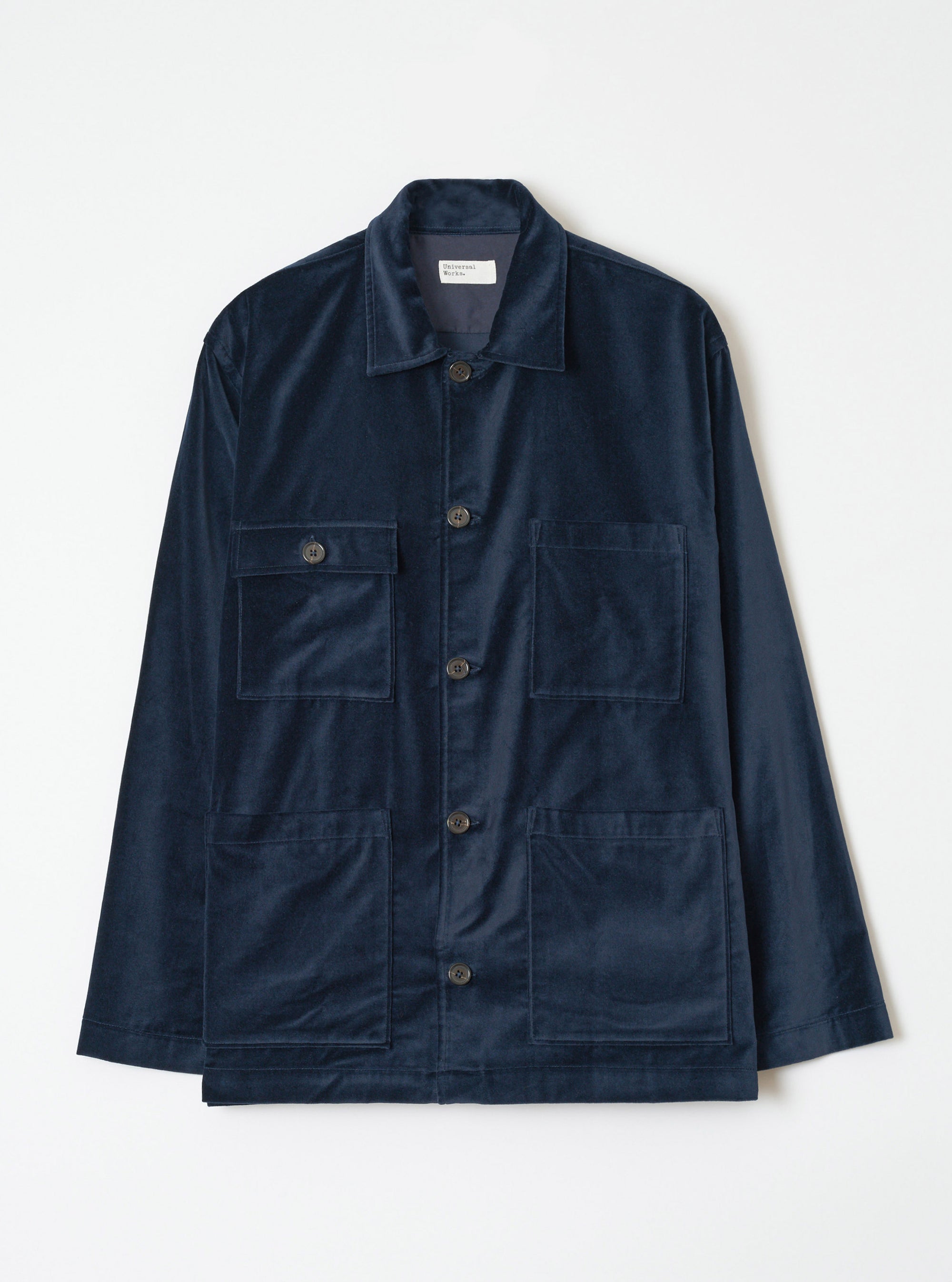 Navy velvet chore jacket laid flat on a white studio backdrop. Features a spread collar, five dark buttons down the placket, two buttoned chest patch pockets and two large lower patch pockets. Long sleeves with plain cuffs. Fabric has a fine, smooth velvet texture. Universal Works label visible at the neck.