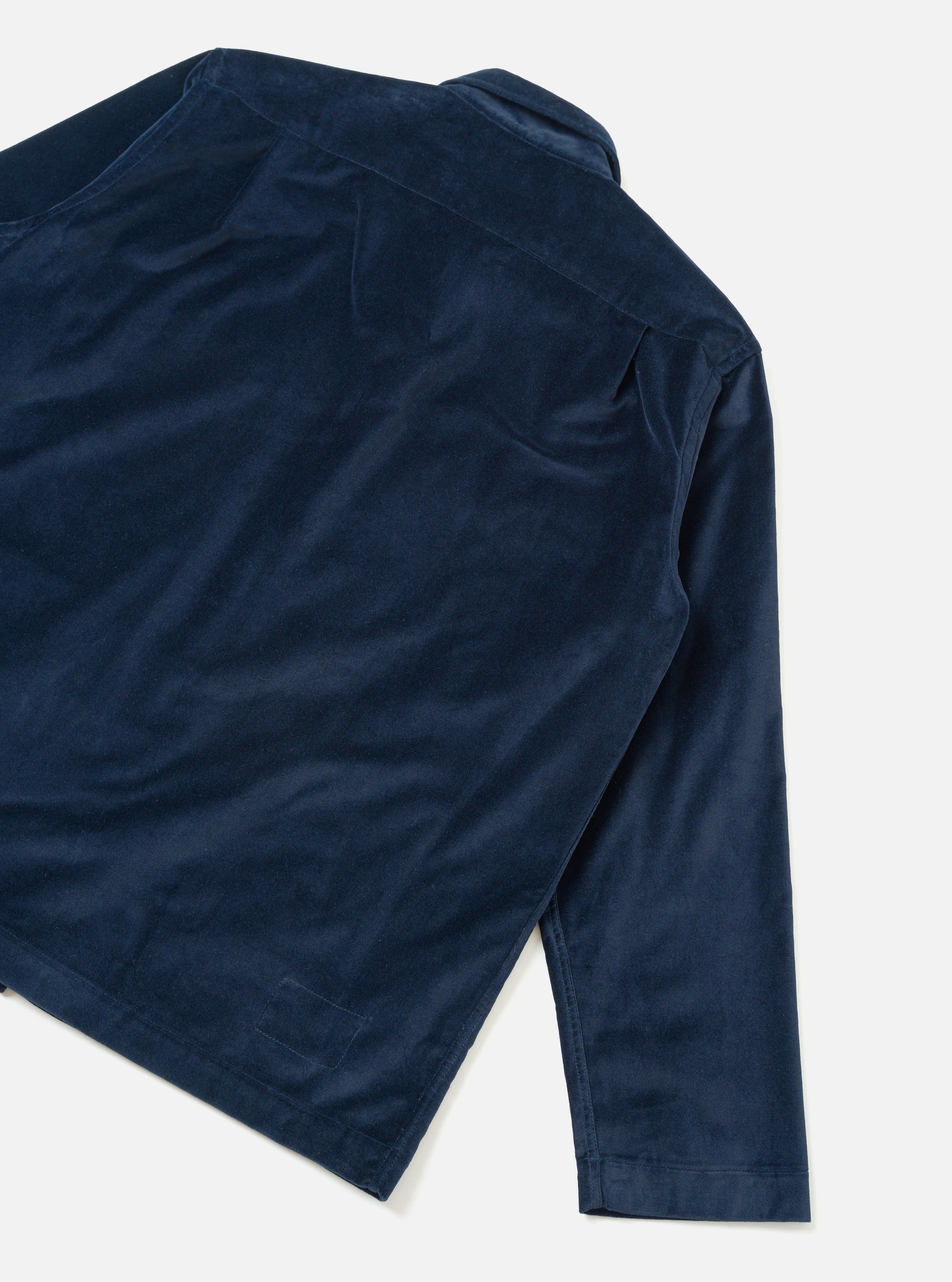 Navy velvet overshirt laid flat on a light grey seamless backdrop, shot from above at an angle. The back and right sleeve are visible, showing the yoke seam, a folded cuff, and the straight hem. The fine-pile velvet fabric catches the light, showing subtle sheen across the body.