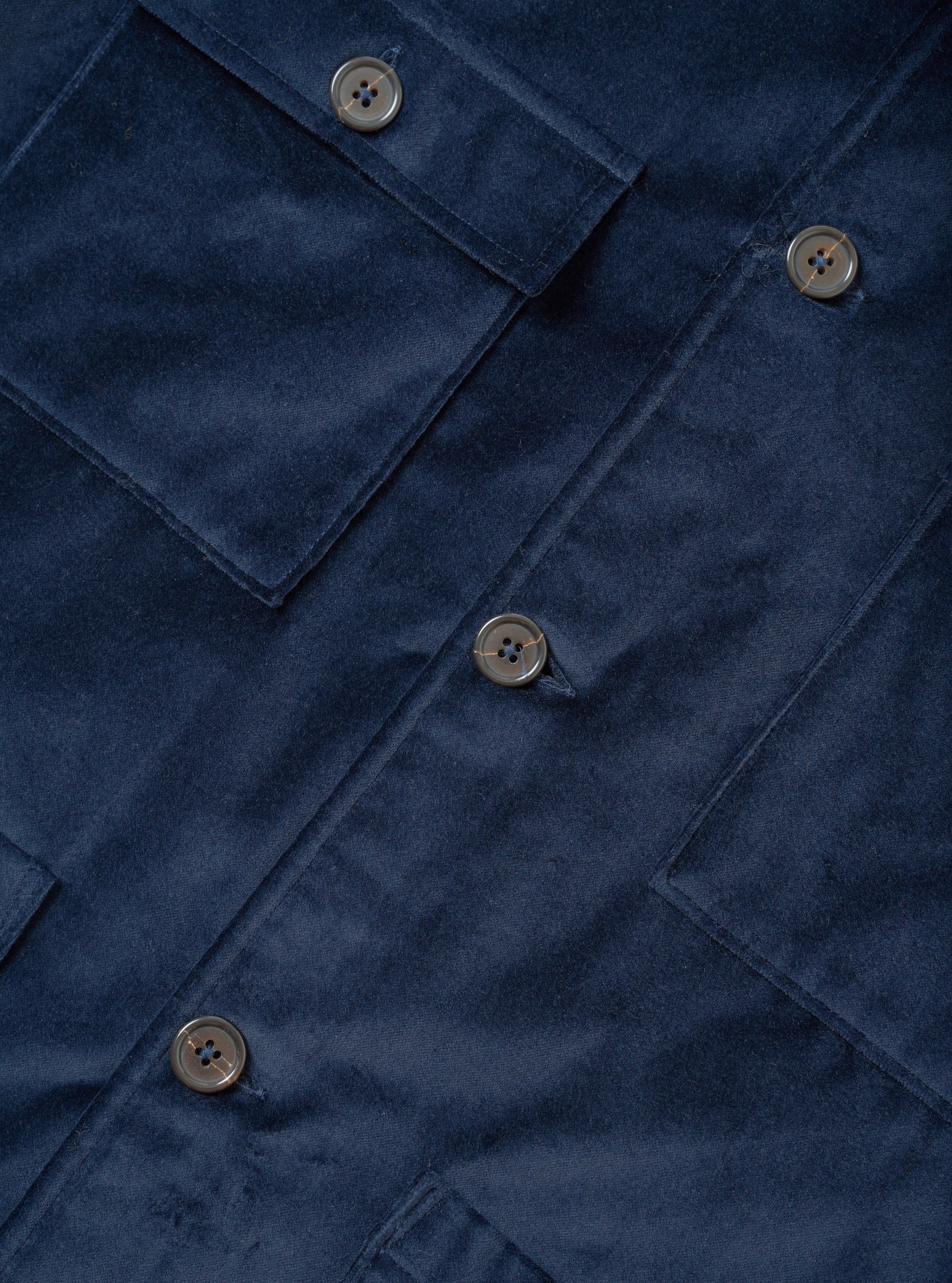 Close-up flat lay of the navy velvet Dockside jacket. The soft, short-pile velvet fabric is shown in detail alongside a buttoned chest patch pocket, a centre-front button placket with four-hole grey buttons, and the edges of two lower patch pockets.