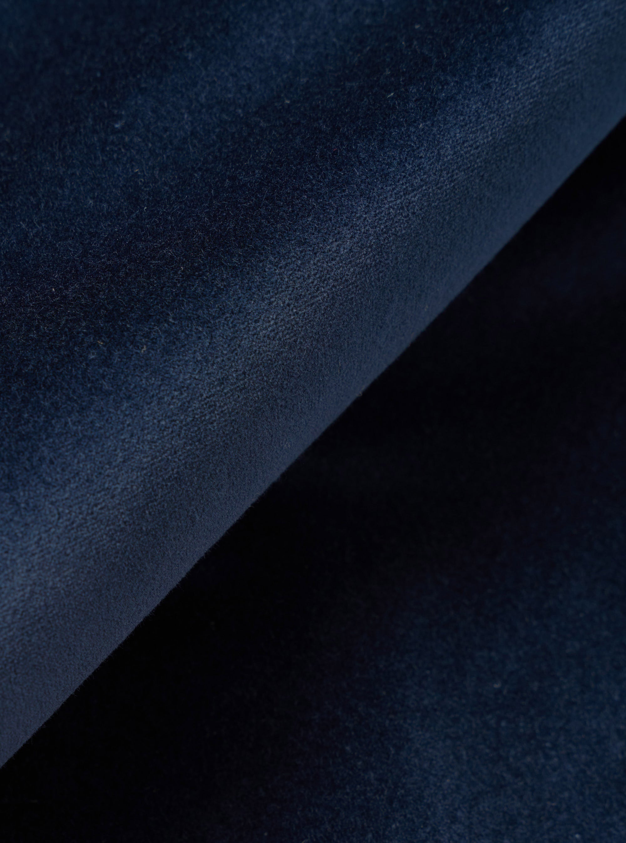 Close-up detail shot of navy velvet fabric, folded diagonally to show the texture and subtle sheen of the pile. The dense, fine-grain surface shows natural variation in depth and tone across the fold. Shot against a dark background.