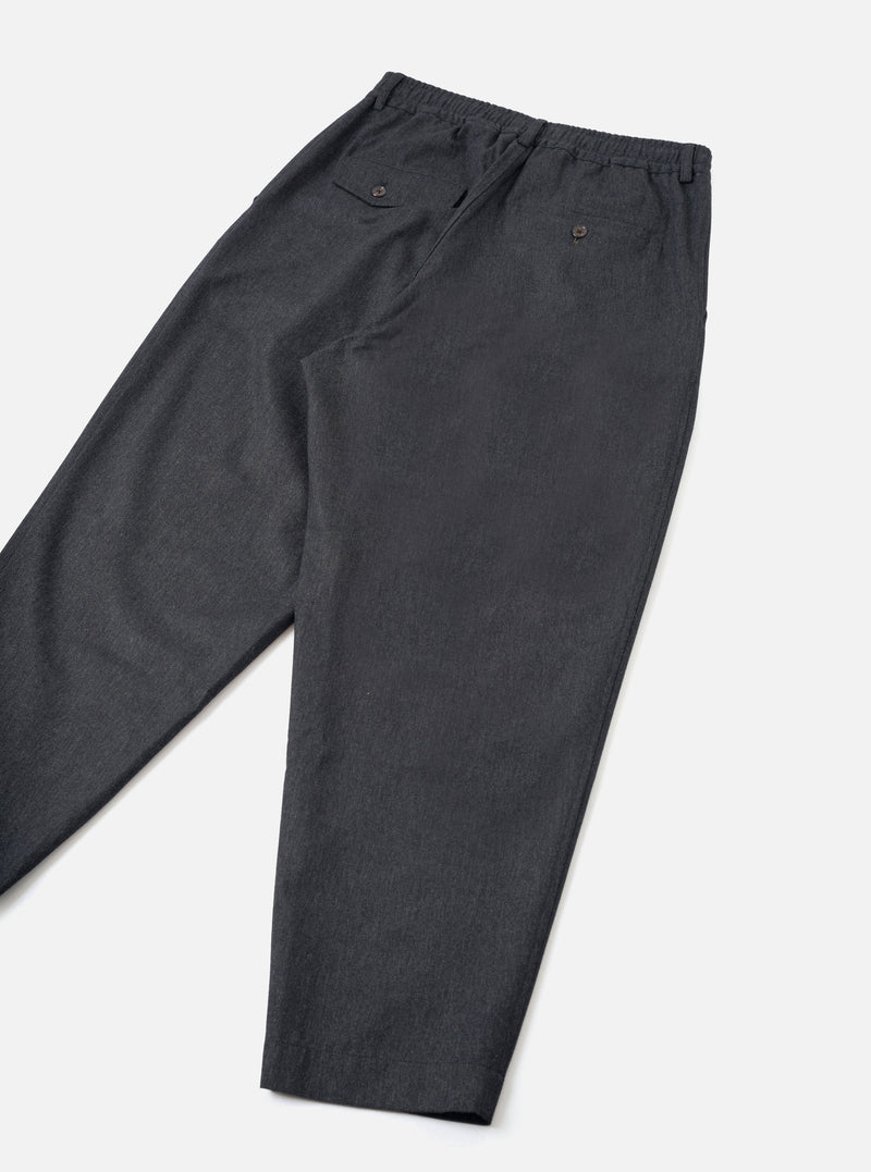A back view of a model wearing the Charcoal Recycled Italian Tweed Pleated Track Pant in a clean studio setting. The image displays the rear pocket placement, comfortable drape, and grey tweed fabric.