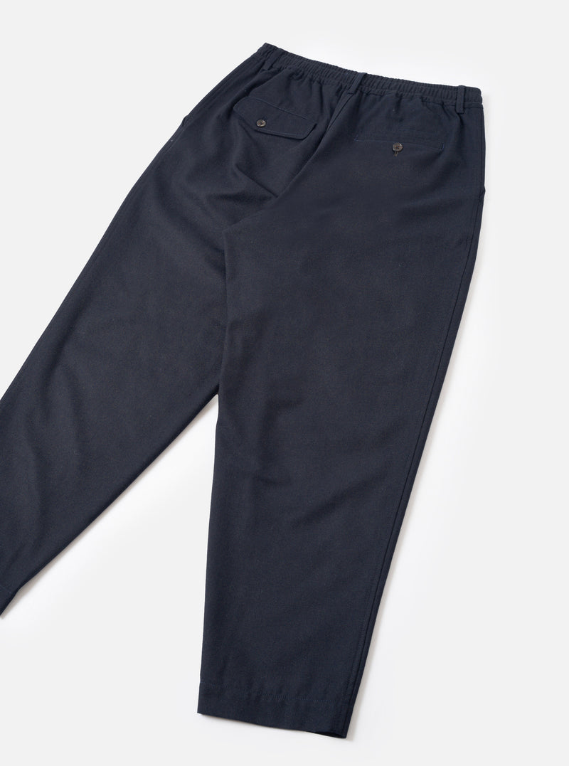 A back view of a model wearing the Navy Recycled Italian Tweed Pleated Track Pant against a neutral studio backdrop. The image shows the relaxed fit, drawstring waistband, and rear patch pocket details of the navy blue trousers.