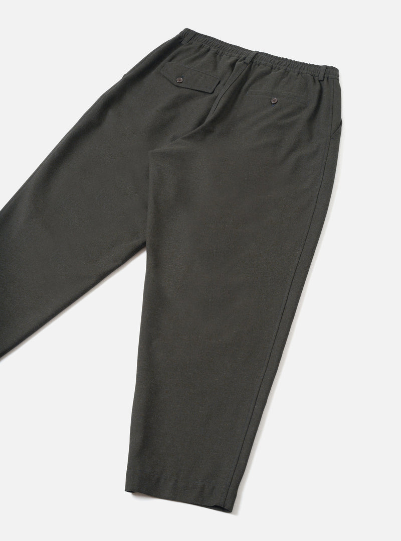 A back view of a model wearing the Olive Recycled Italian Tweed Pleated Track Pant against a neutral studio backdrop. The photograph showcases the clean hem, rear pocket detail, and comfortable fit of the olive green trousers.
