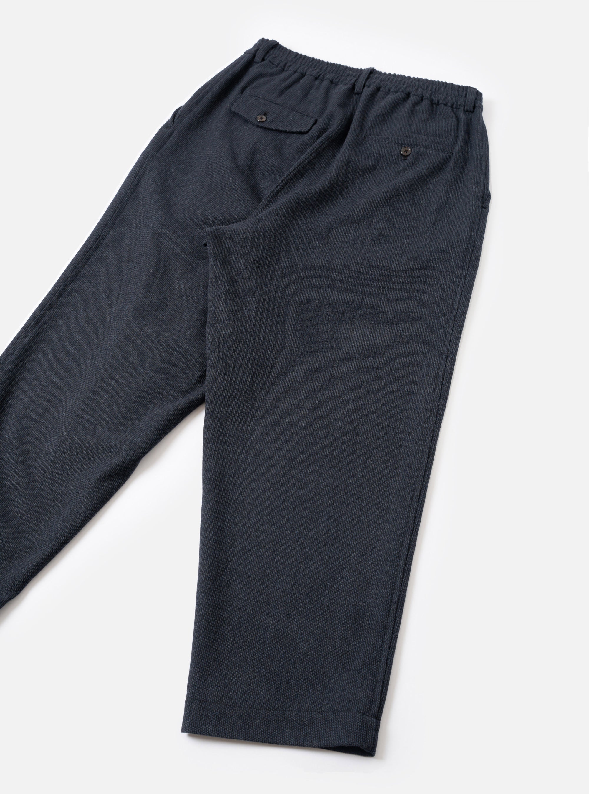 Navy flannel oxford trousers laid flat on a white surface, showing the rear. The elasticated waistband is visible at the top, with two back pockets: one buttoned flap pocket on the left and one button-through welt pocket on the right. The fabric has a fine textured weave. The tapered legs end in turn-up cuffs.