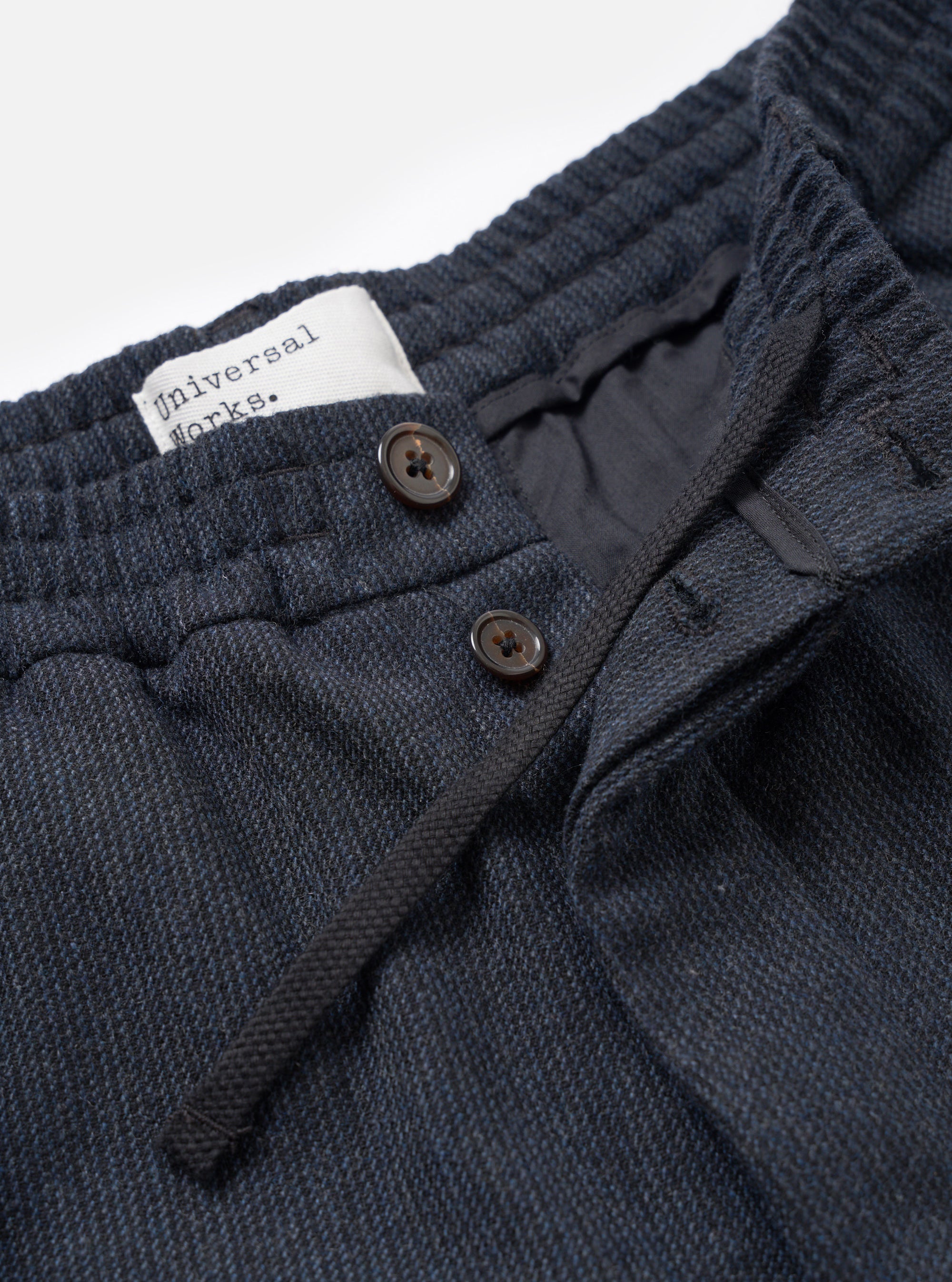 Close-up of the waistband detail on the Universal Works navy flannel Oxford pant. A wide elasticated waistband sits above a button fly with two dark horn-style buttons and a woven drawcord. A white branded label is visible inside. The twill-weave flannel fabric shows a navy and dark grey marl texture. Plain white studio backdrop.