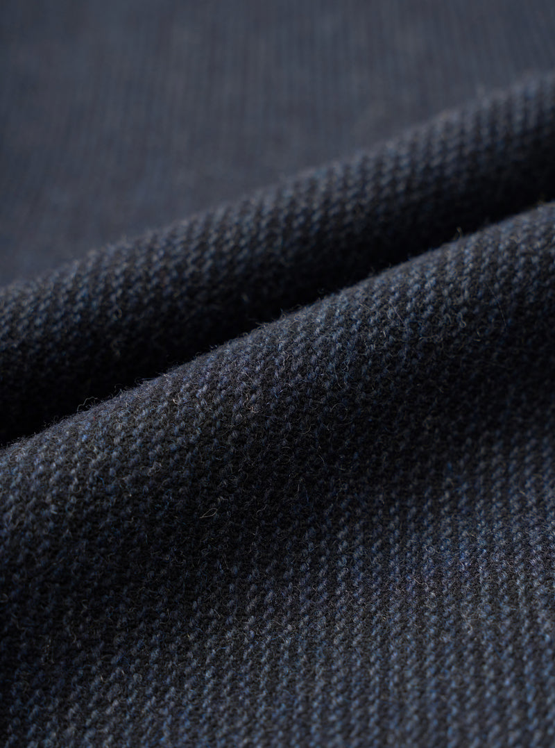 Close-up fabric detail of the navy airline flannel oxford pant, showing a charcoal and navy twill-weave wool flannel with a fine diagonal rib texture and visible surface fibres. The fabric is folded to reveal depth and drape, shot against a matching dark flannel background.