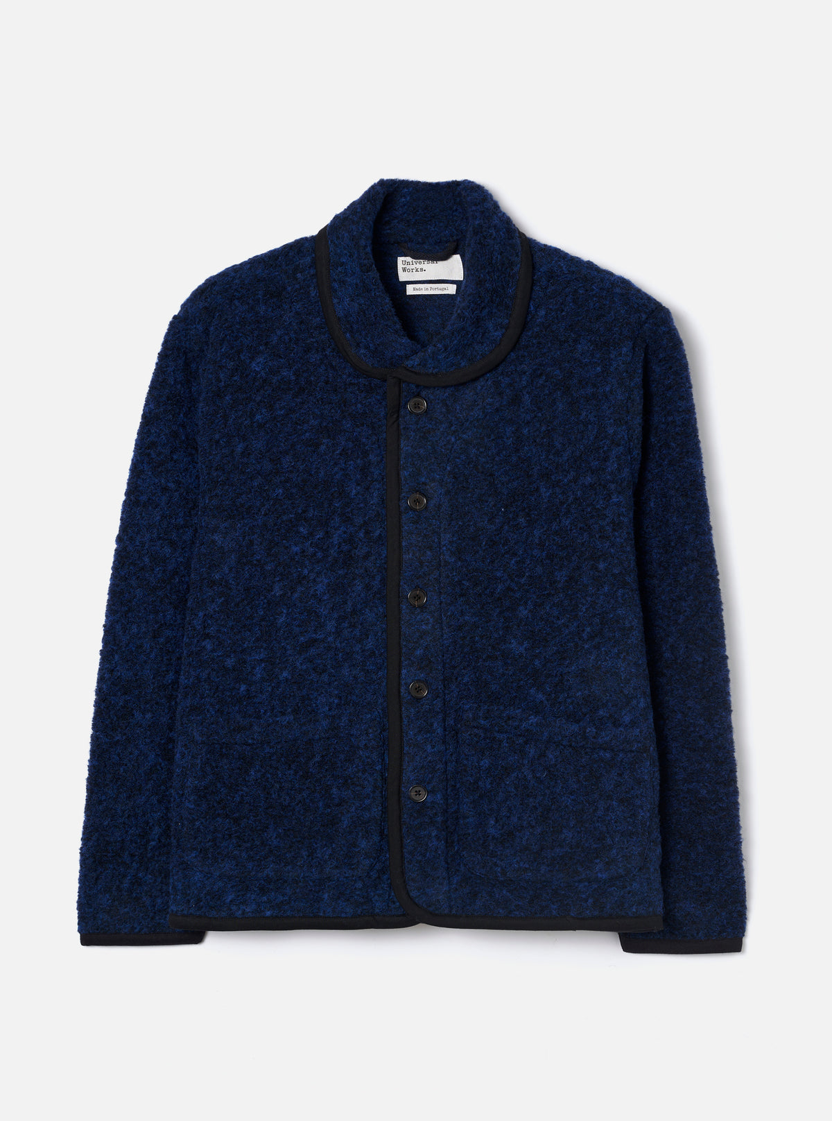 Blue boucle style fleece Lancaster jacket laid flat on a light grey seamless backdrop. The jacket features a shawl collar, five dark buttons down the centre front, and a patch pocket at the lower left. Black binding trims the collar, front placket, cuffs, and hem. A Universal Works branded label is visible at the inner collar.