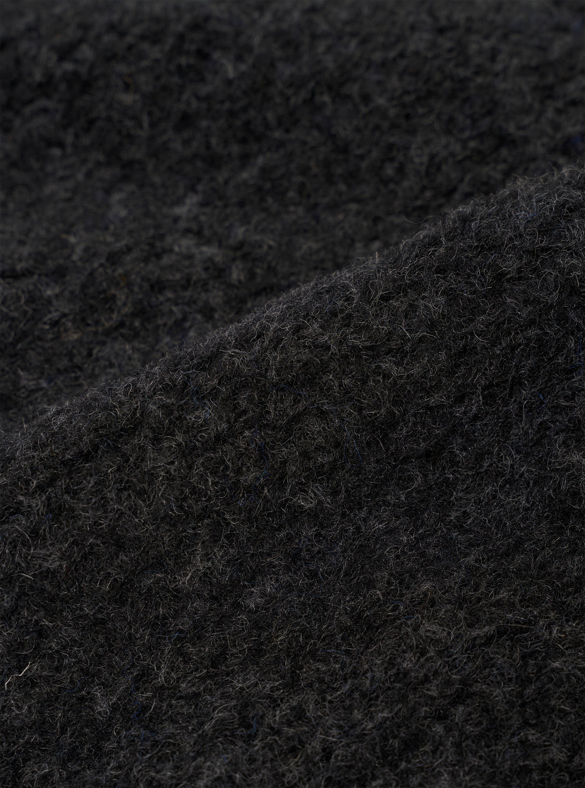 Close-up detail of the charcoal boucle style fleece fabric used on the Lancaster Jacket. The shot shows the dense, curly looped texture of the boucle style weave in dark charcoal grey, with individual fibres visible across the surface. A folded edge runs diagonally across the frame, highlighting the fabric's lofty, textured depth.