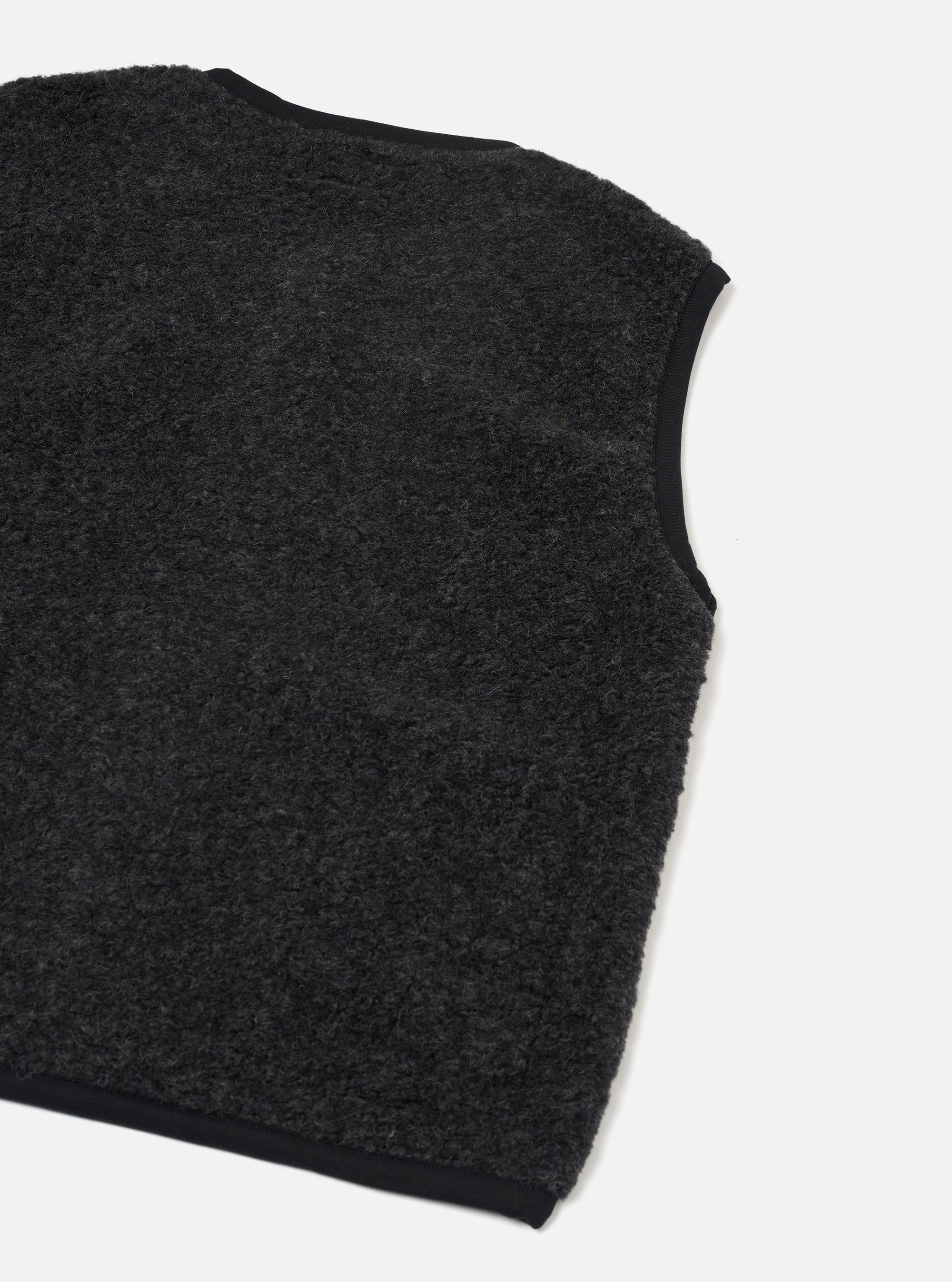 Charcoal boucle style fleece gilet laid flat on a white studio surface, shot at an angle showing the back and one armhole. The textured, looped fleece body is charcoal grey with contrast black binding along the armhole edge, neckline and hem.