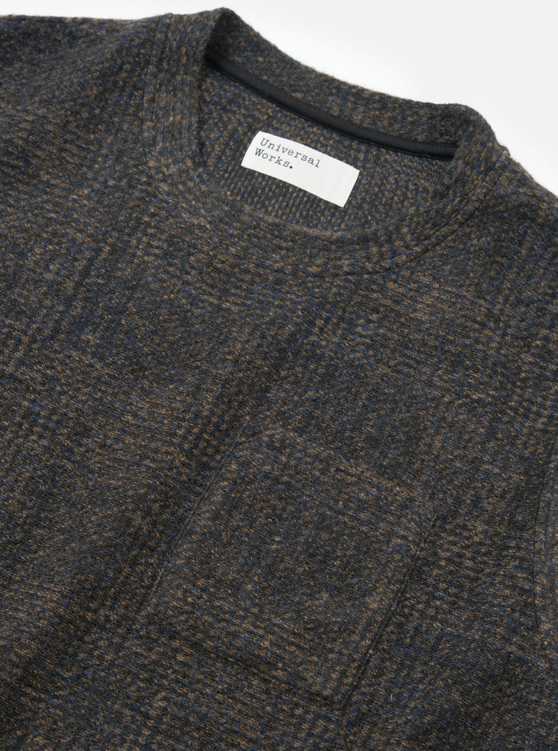 Close-up flat lay of a Universal Works navy Deca Light Knit Sleeveless Crew on a white studio surface. The marl fabric blends navy, brown and blue tones in a fine knit texture. A ribbed crew neckline is visible with a dark inner tape, and a woven Universal Works label sits at the front neck.